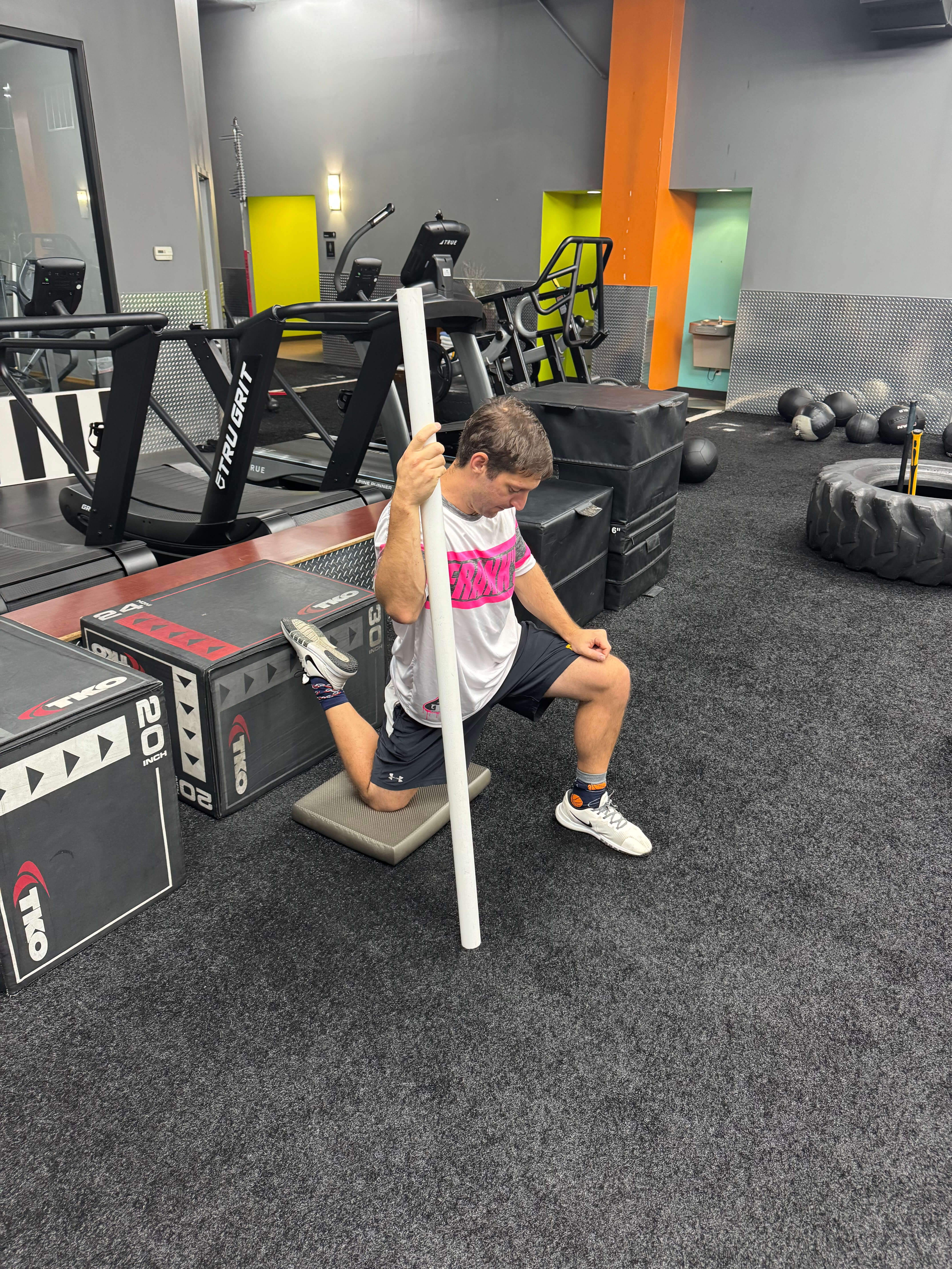 Man in a gym kneeling on a padded mat while holding a white pole for support during a stretching exercise.