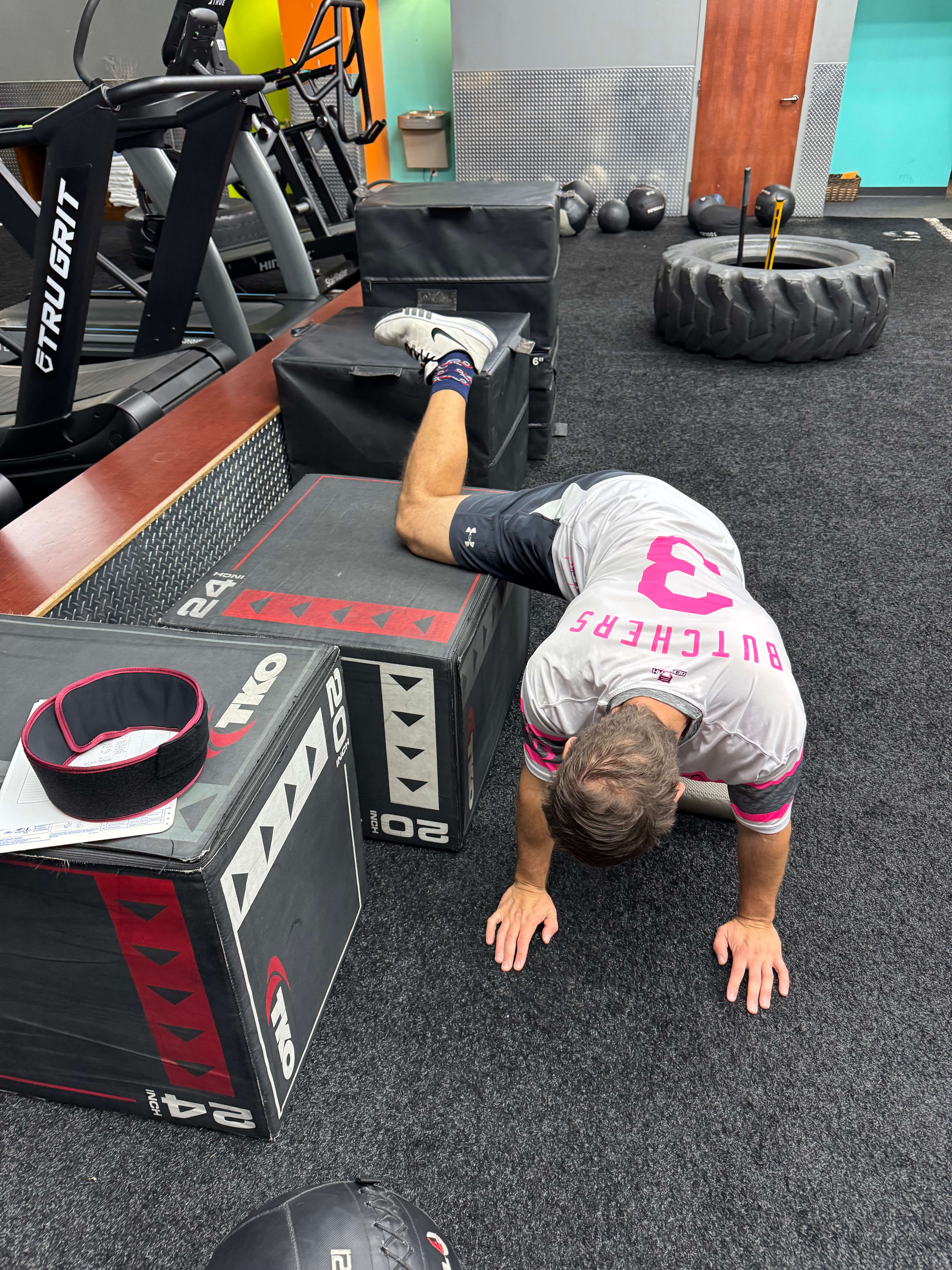 Man performing a stretching exercise with one leg bent and resting on a padded box in a gym setting.
