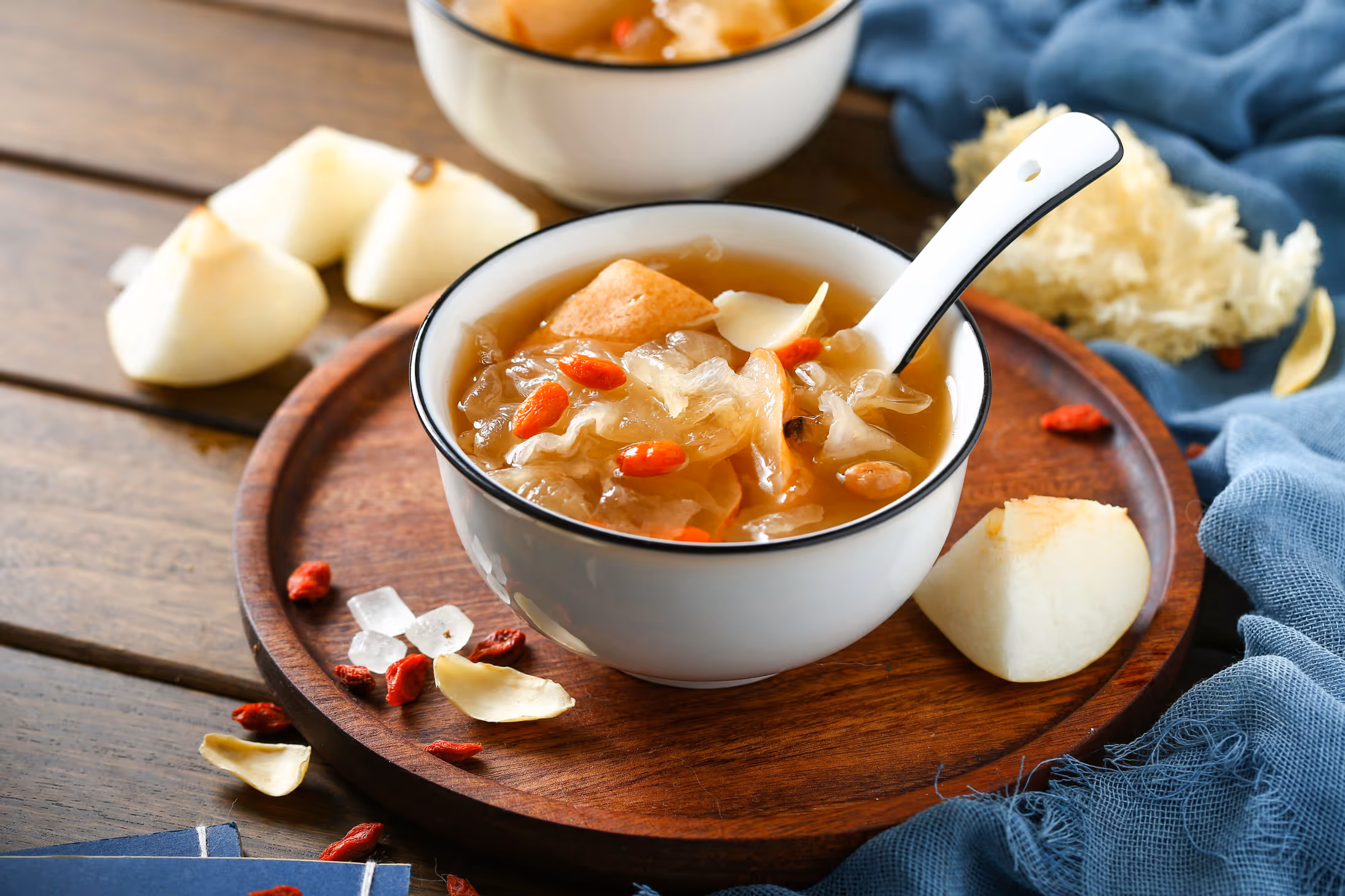 White ceramic bowl with a spoon containing Asian dessert soup with white fungus, goji berries, and pear slices on a wooden tray.
