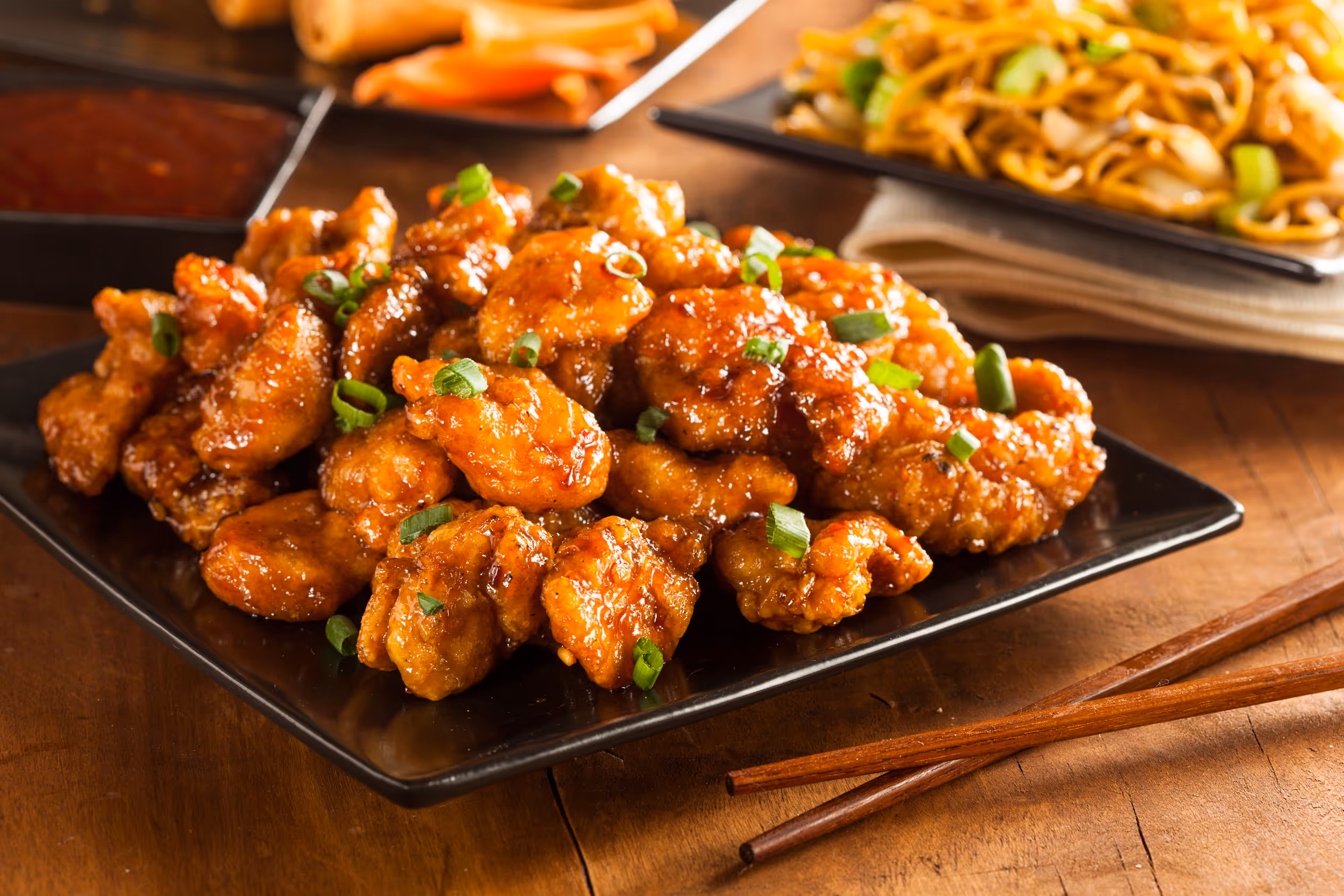 Plate of crispy orange chicken garnished with chopped green onions on a wooden table with chopsticks.