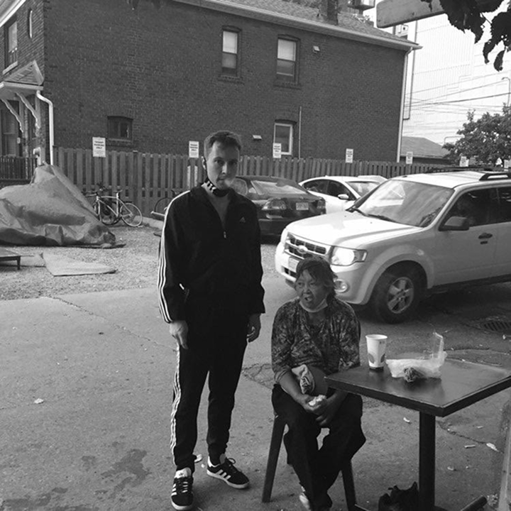 Man in tracksuit standing beside woman seated at table outdoors on urban street with parked cars.