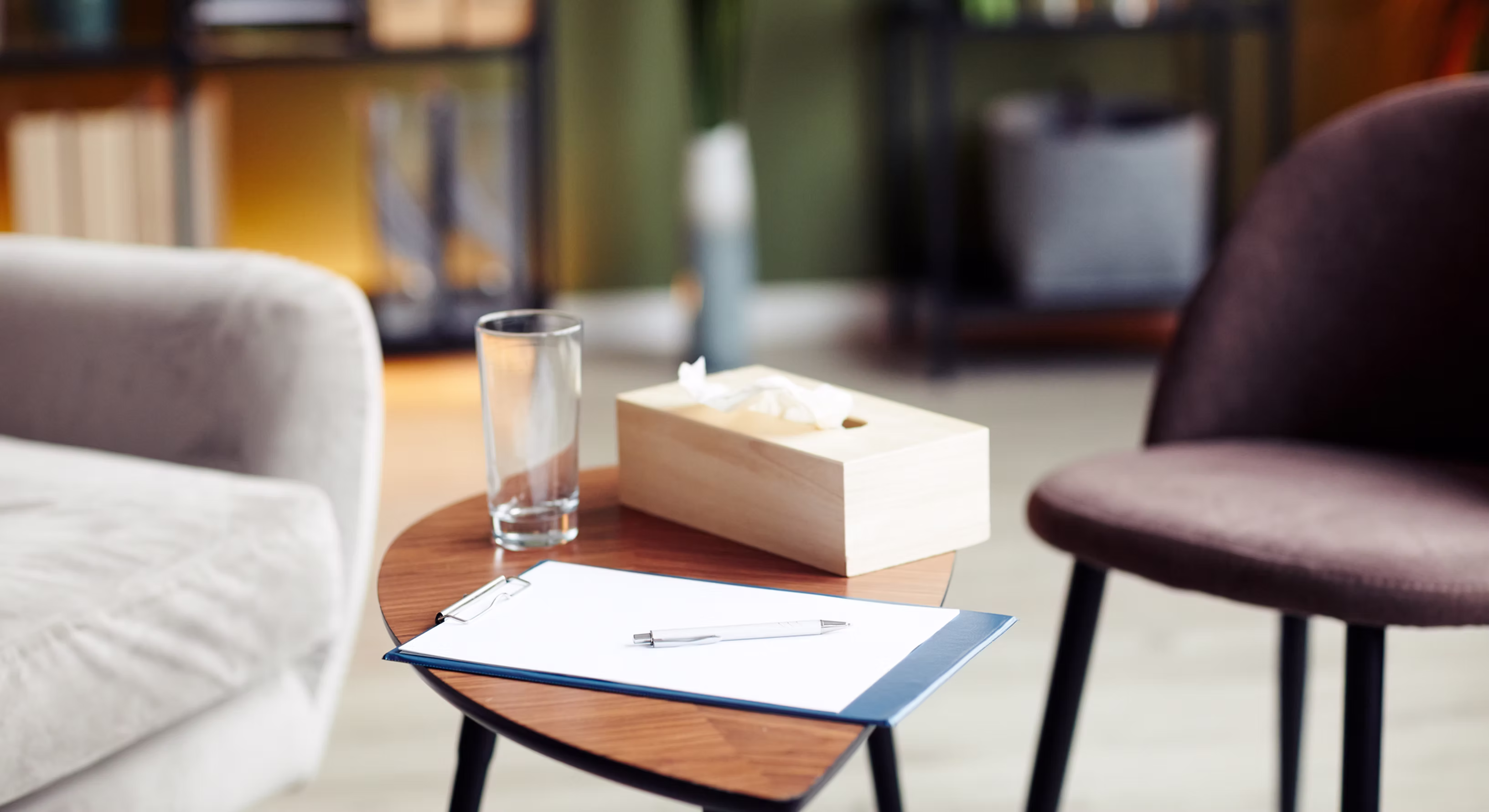 A calm therapy office with two chairs, a clipboard, a glass of water, and a tissue box on a side table.