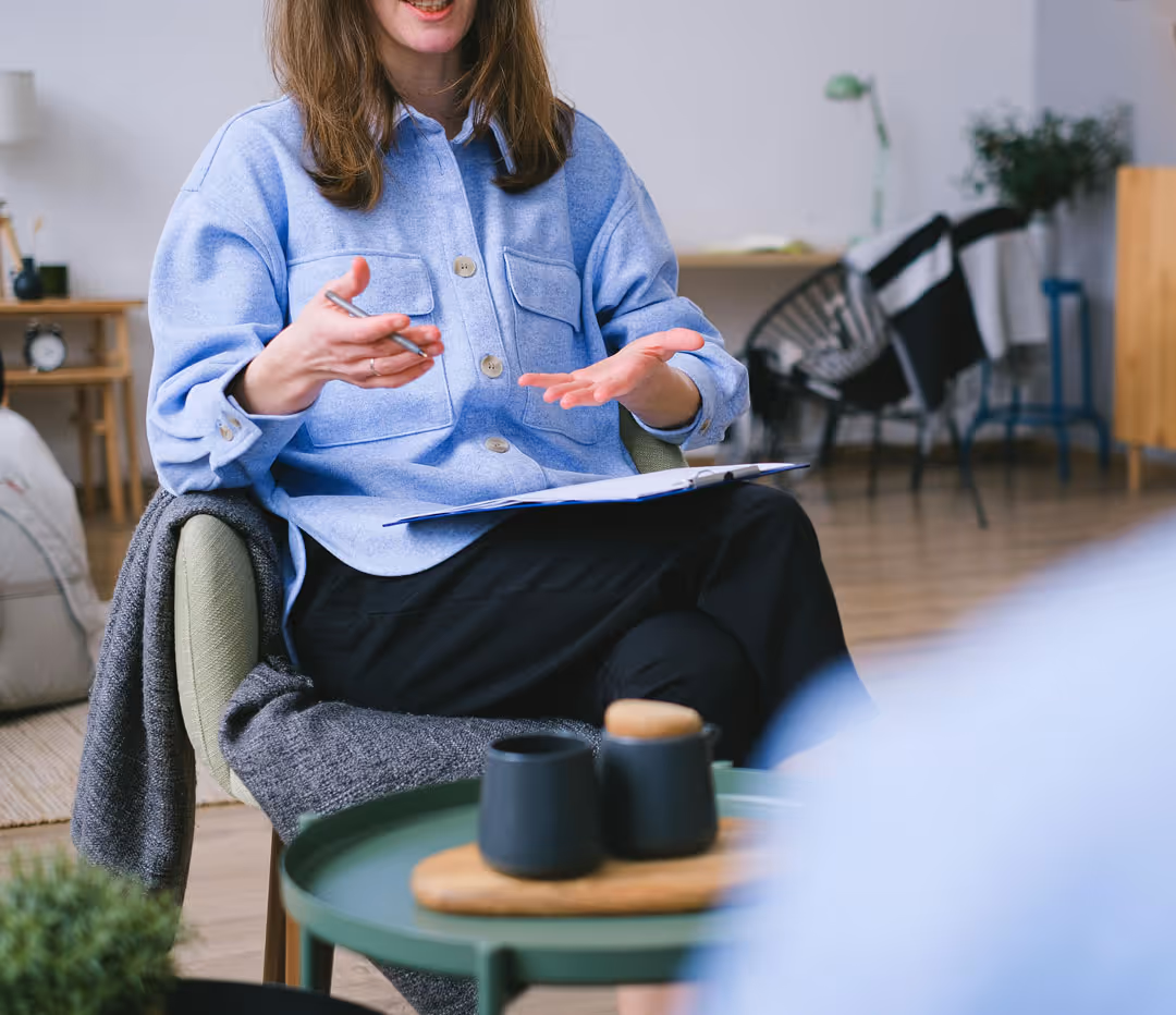 A therapist sits with a clipboard and speaks warmly with a client during a therapy session in a cozy office setting.