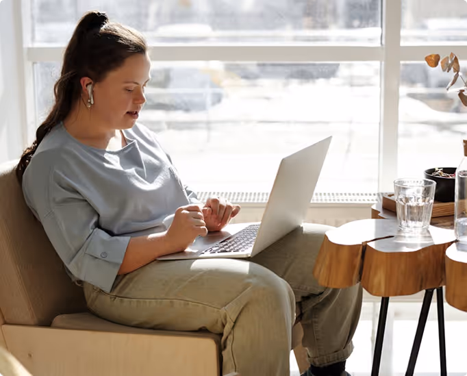 A young woman with Down syndrome sits comfortably in a bright, modern space, working on her laptop with wireless earbuds in.