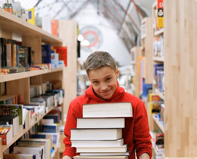 A smiling child carries a tall stack of books through a school library, representing a love of learning and educational support.