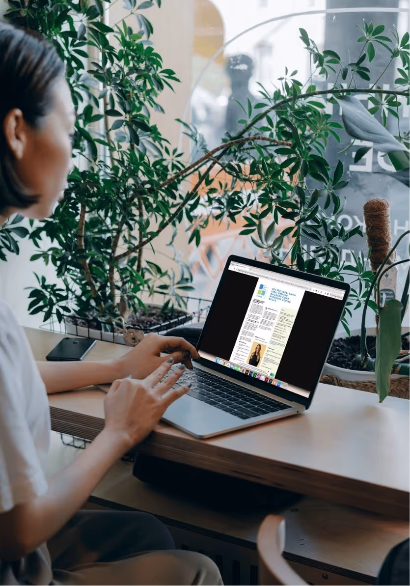 A woman sits at a wooden desk surrounded by lush green plants, reading the Little Tree Psychology Corner newsletter on her laptop.