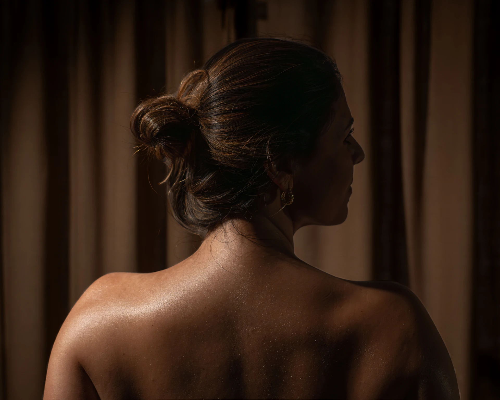Mujer de espaldas con cabello recogido en un moño y aretes dorados frente a cortinas marrones.