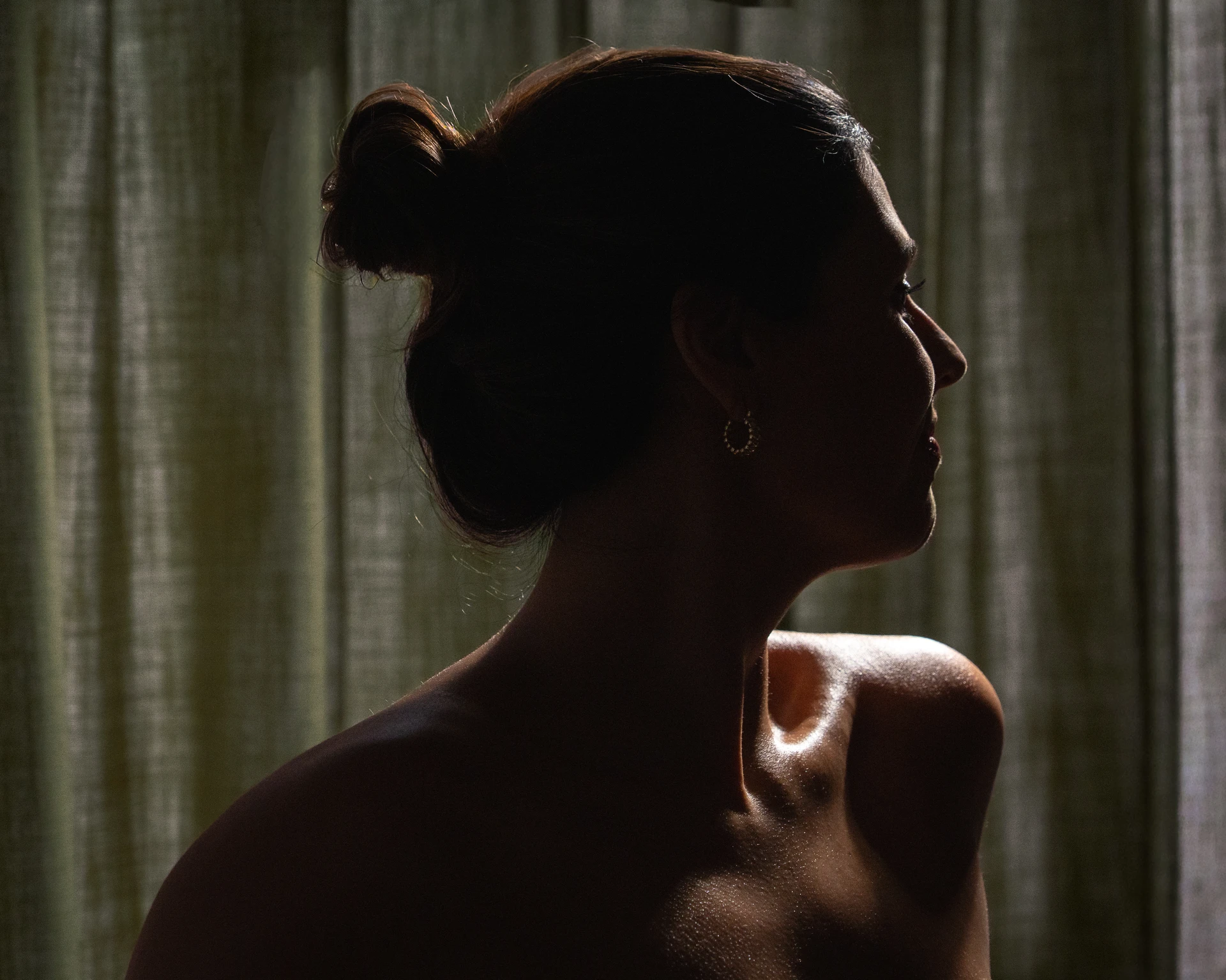 Perfil de una mujer con cabello recogido en un moño y pendiente de aro, iluminada suavemente contra una cortina.