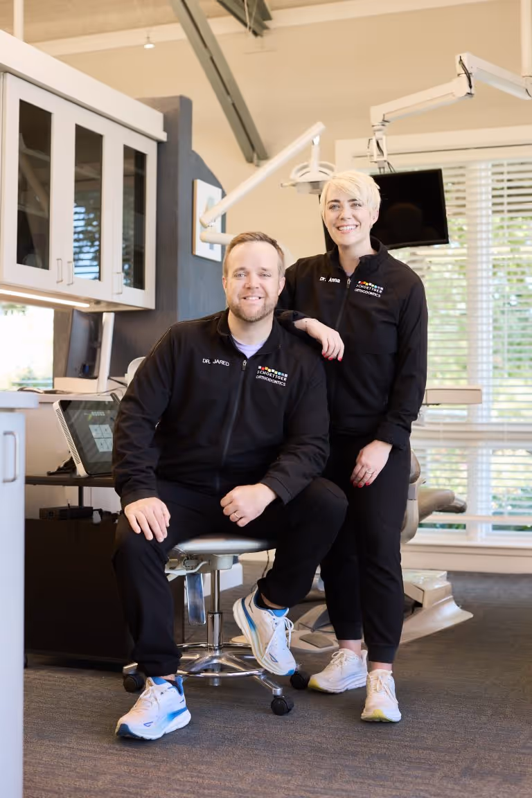 Two orthodontists in black jackets with 'Schoettger' logo posing in a dental office, one seated and one standing.