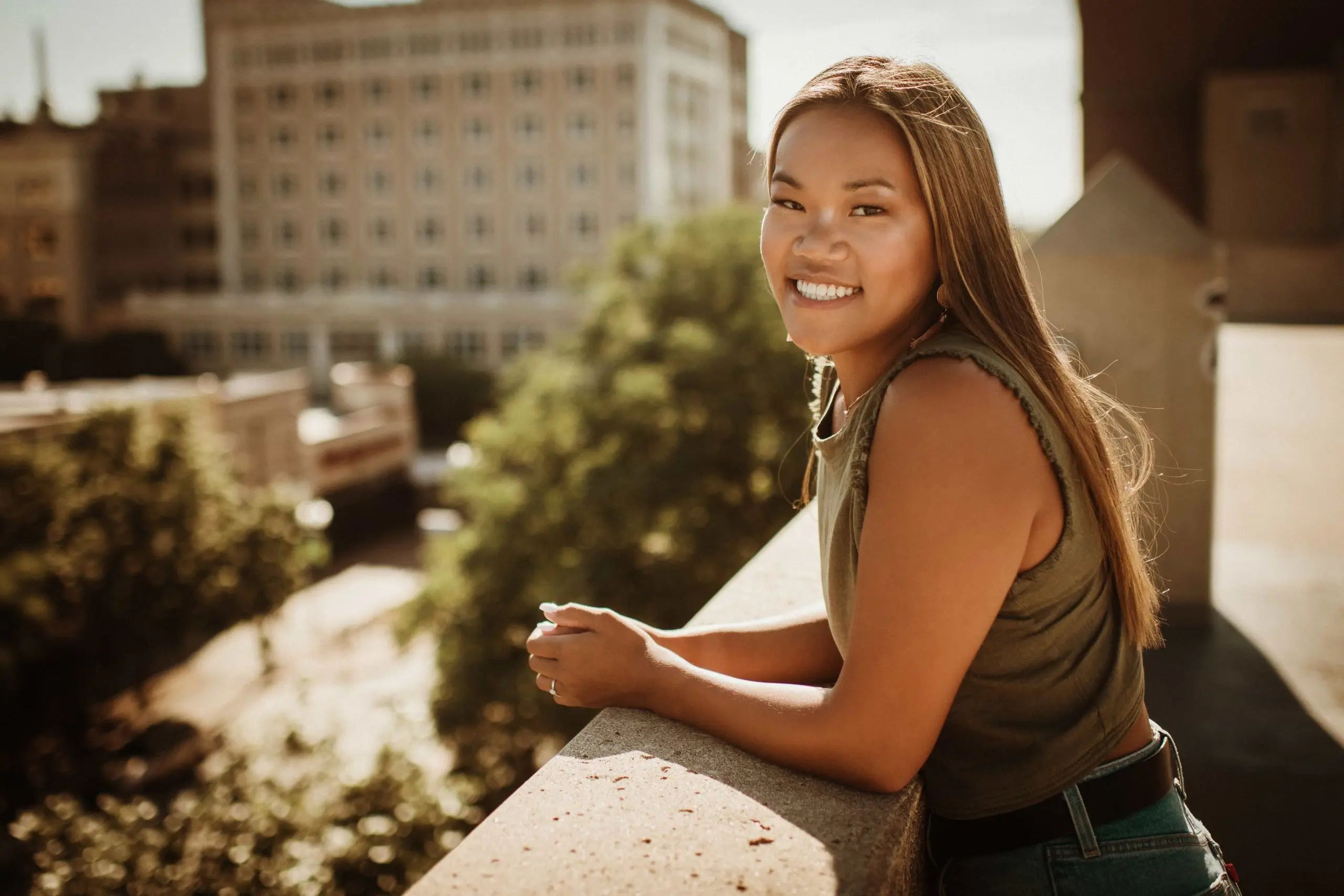 Smiling young woman with long hair leaning on a concrete balcony railing with blurred city buildings and greenery in the background.