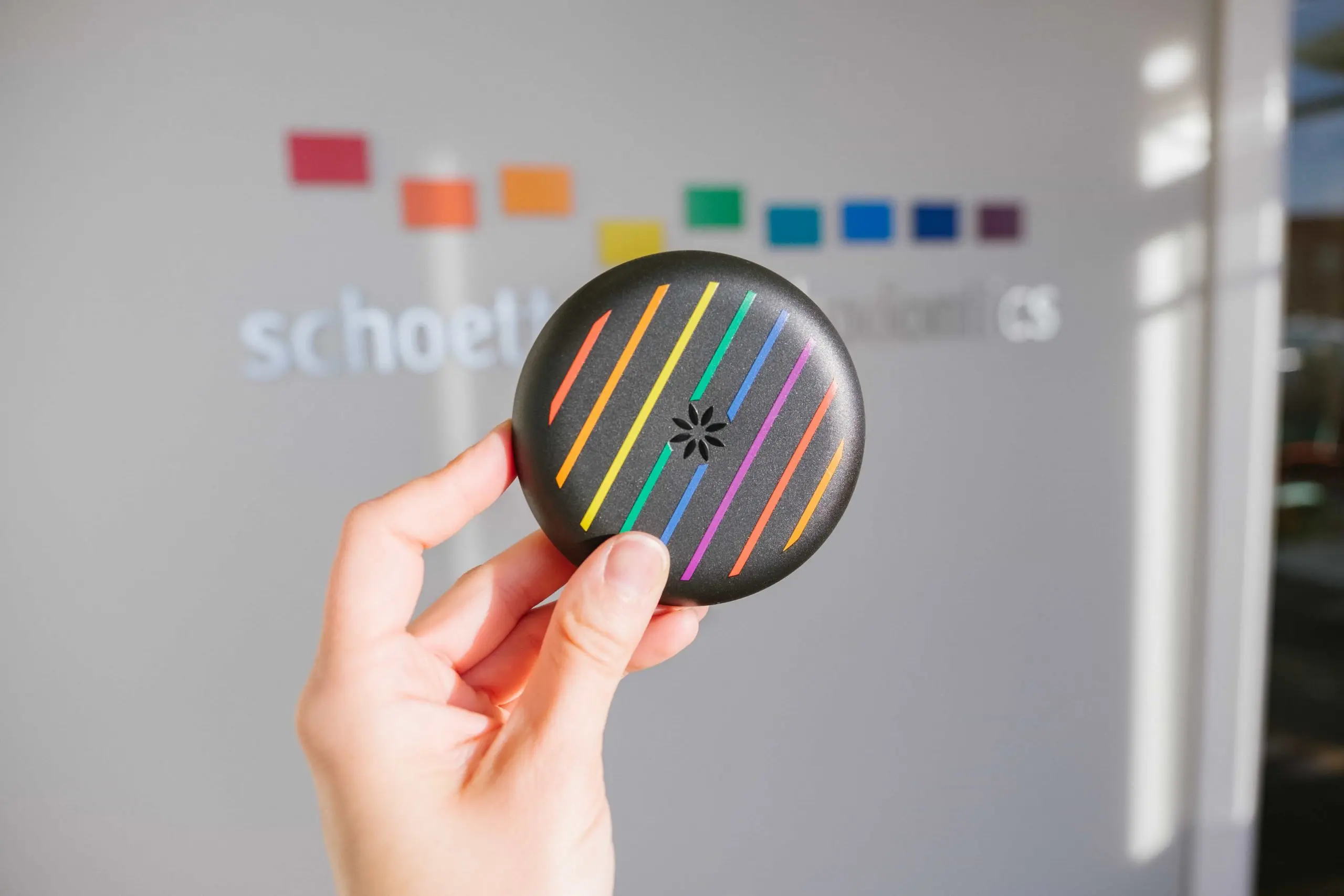 Hand holding a black round object with colorful diagonal stripes and a small flower-like cutout in the center.