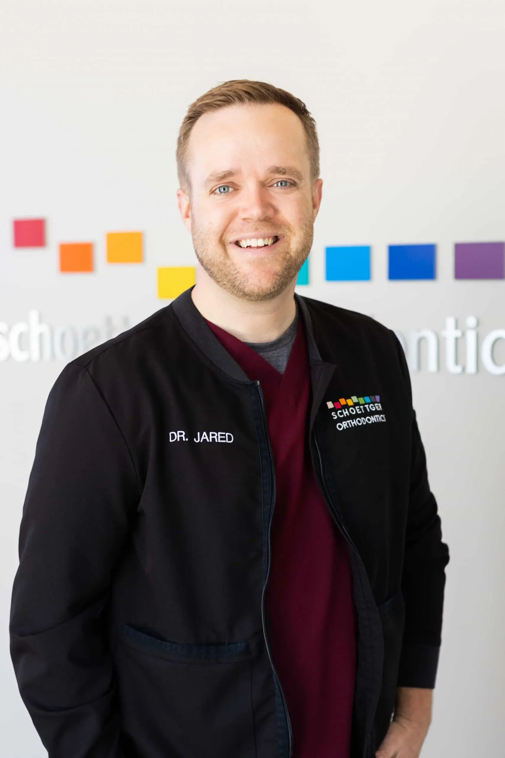 Smiling man wearing a black jacket embroidered with 'Dr. Jared' and 'Schoettger Orthodontics' standing in front of a wall with colorful squares.
