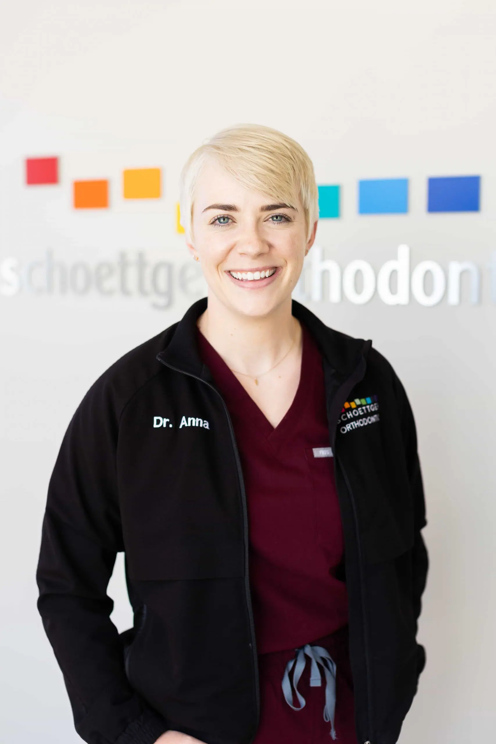 Smiling woman with short blonde hair wearing a maroon scrub top and black jacket embroidered with 'Dr. Anna' and orthodontics logo.