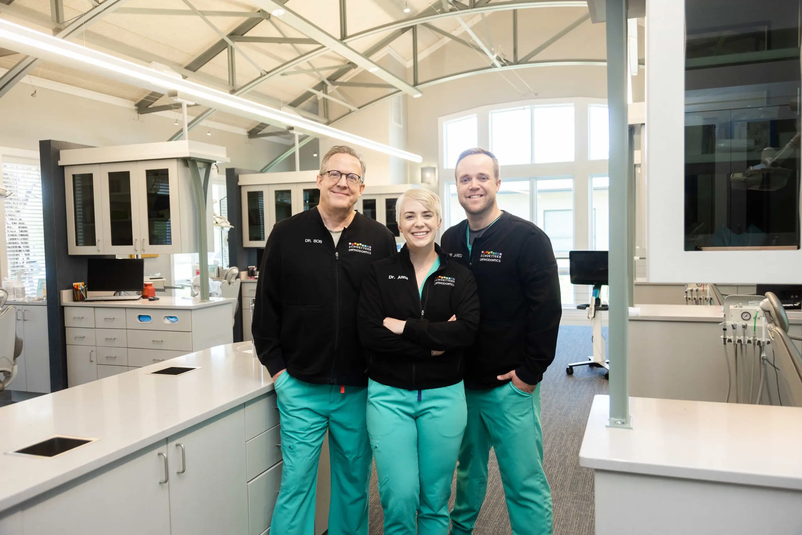 Three dental professionals in black jackets and teal scrubs standing together and smiling inside a modern orthodontic clinic.