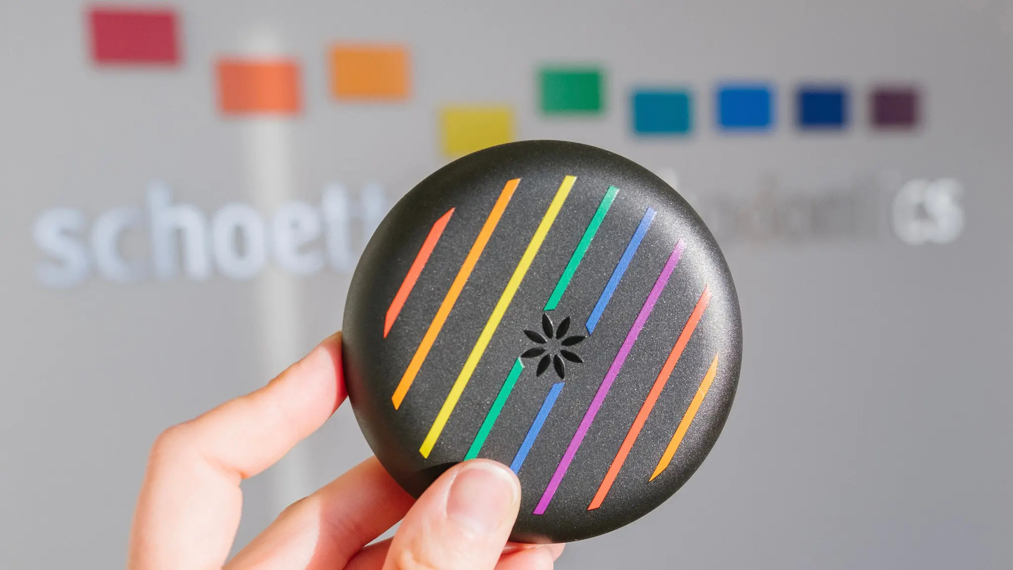 Hand holding a round black device with diagonal rainbow-colored stripes and a flower-shaped vent in the center.