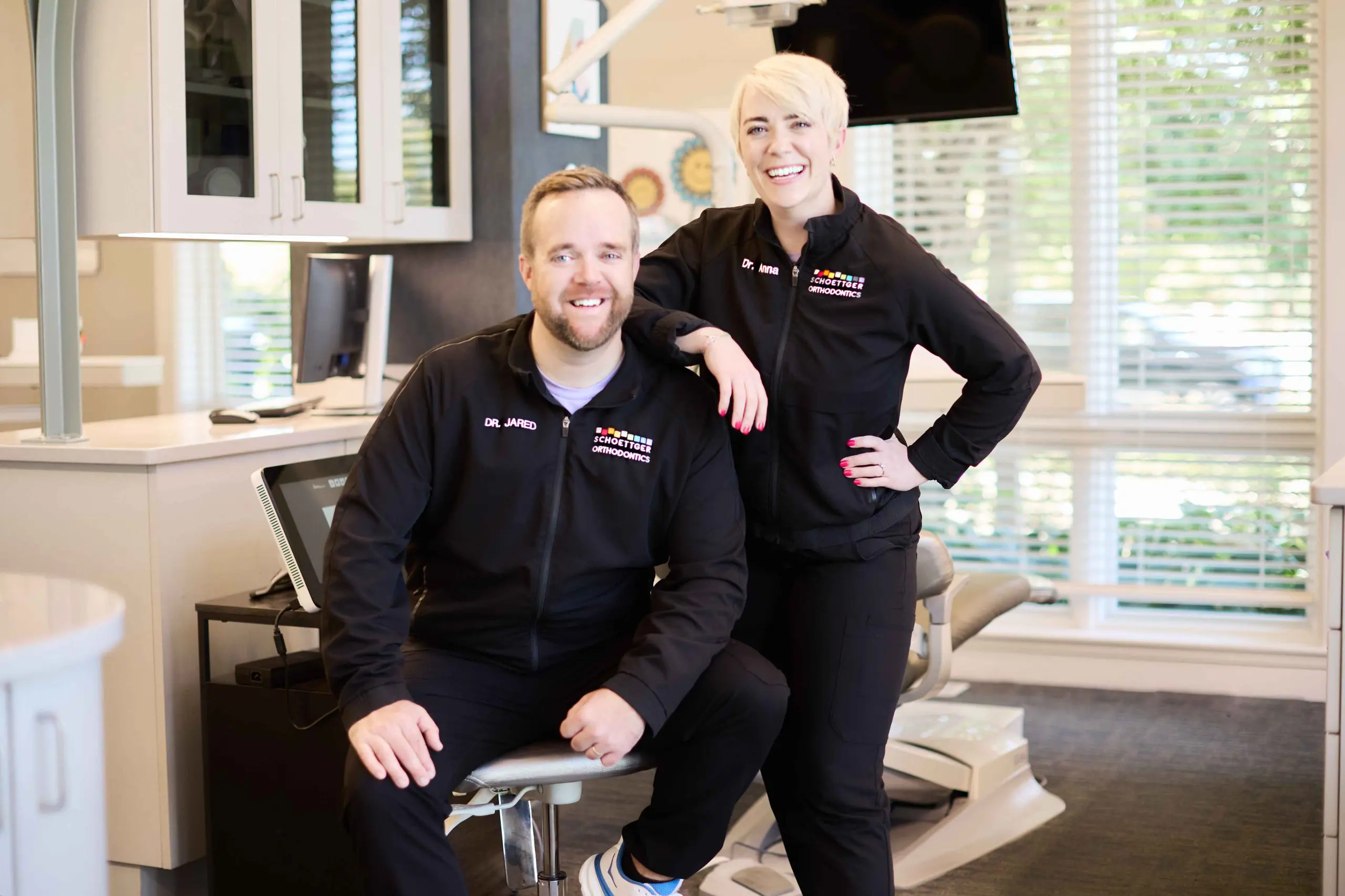 Two smiling orthodontists in black jackets labeled 'Dr. Jared' and 'Dr. Anna' posing in a bright orthodontic office.
