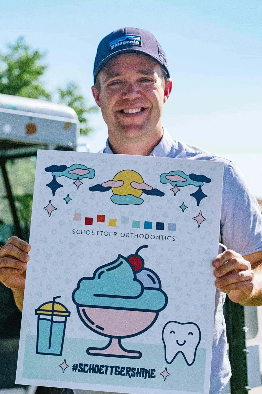 Smiling man wearing a Patagonia hat holding a Schoettger Orthodontics sign with colorful illustrations of a sundae, a drink, a smiling tooth, and the hashtag #SCHOETTGER.SHINE.