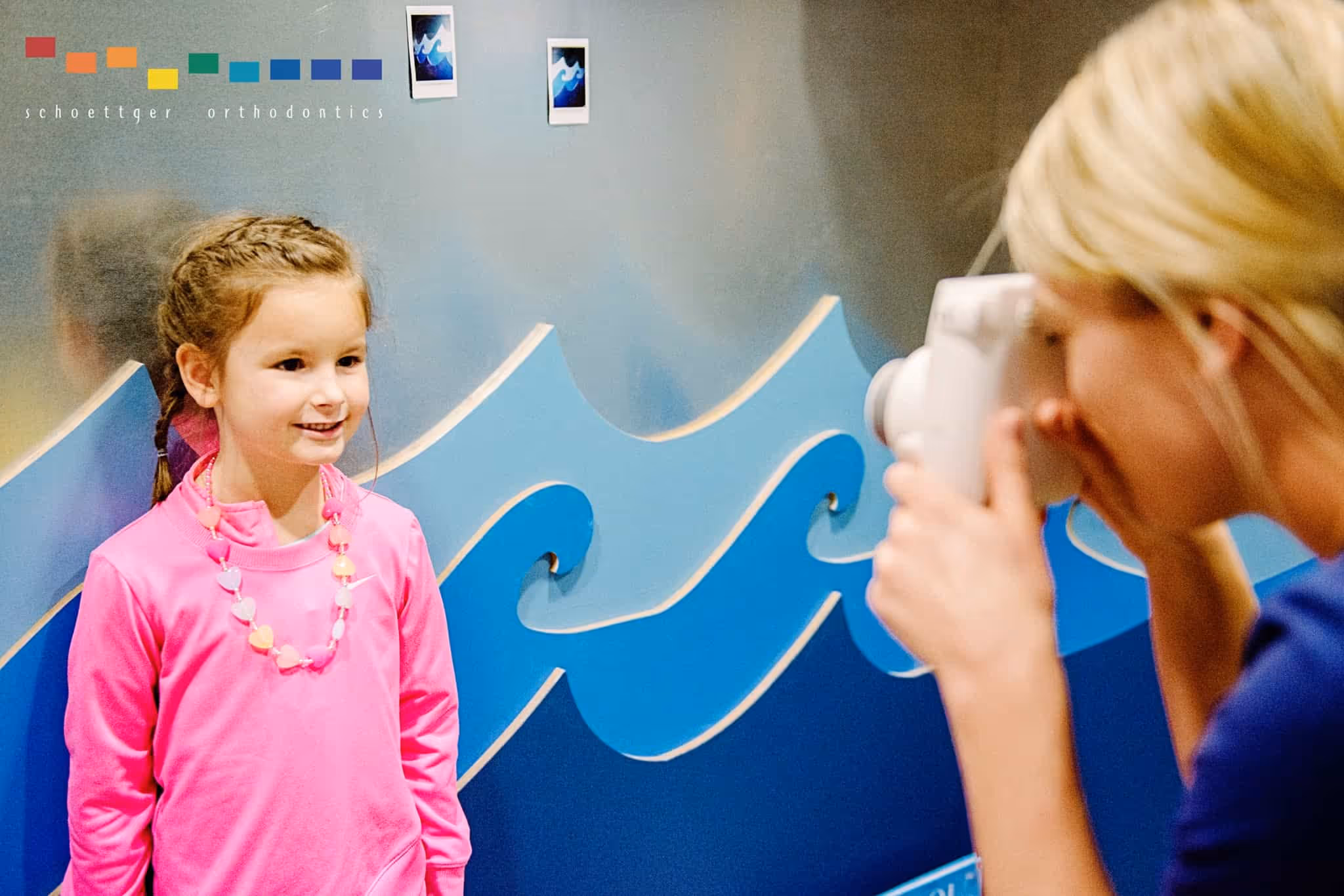 Young girl in a pink shirt smiling as a woman takes her photo with an instant camera against a blue wave-patterned wall.