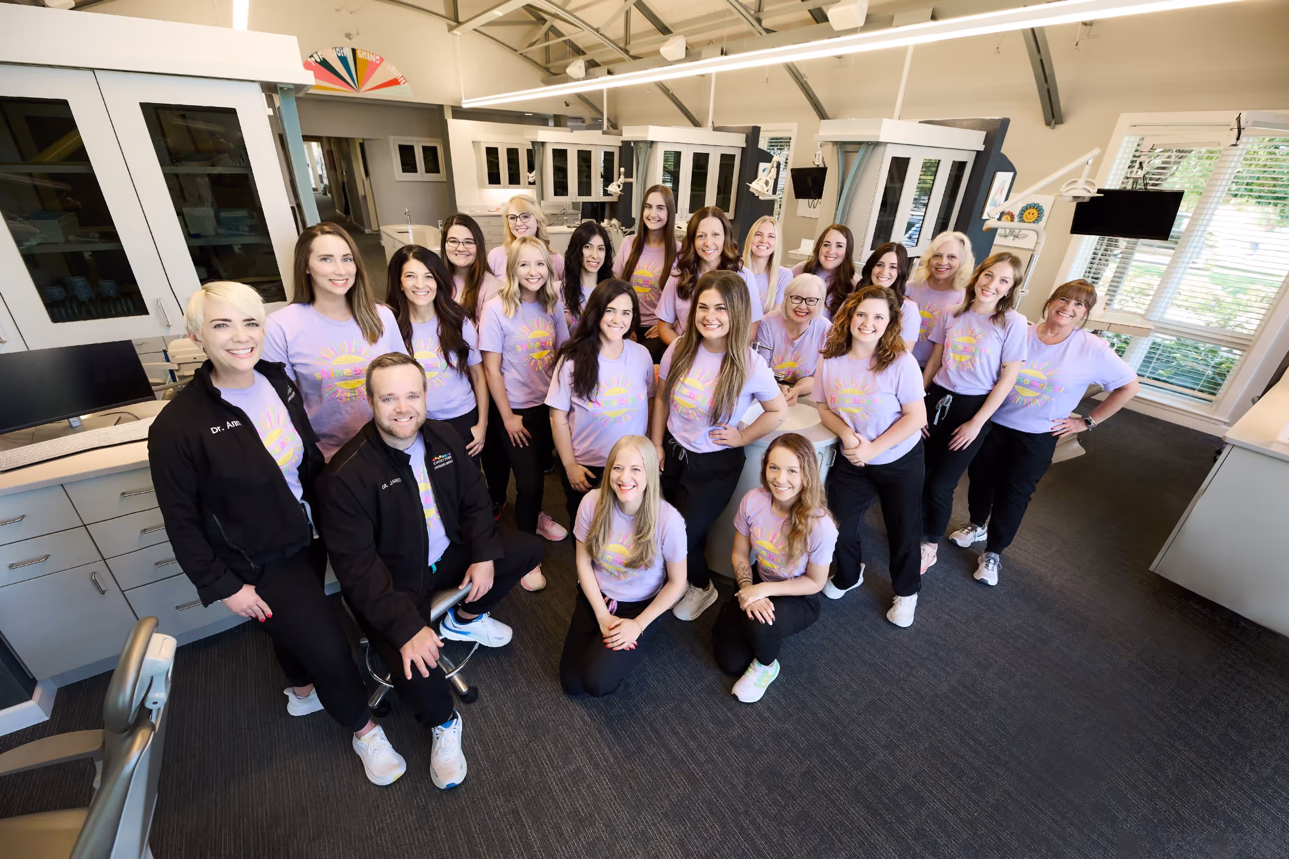 Group of smiling dental office staff wearing matching purple t-shirts posing inside a modern orthodontic clinic.