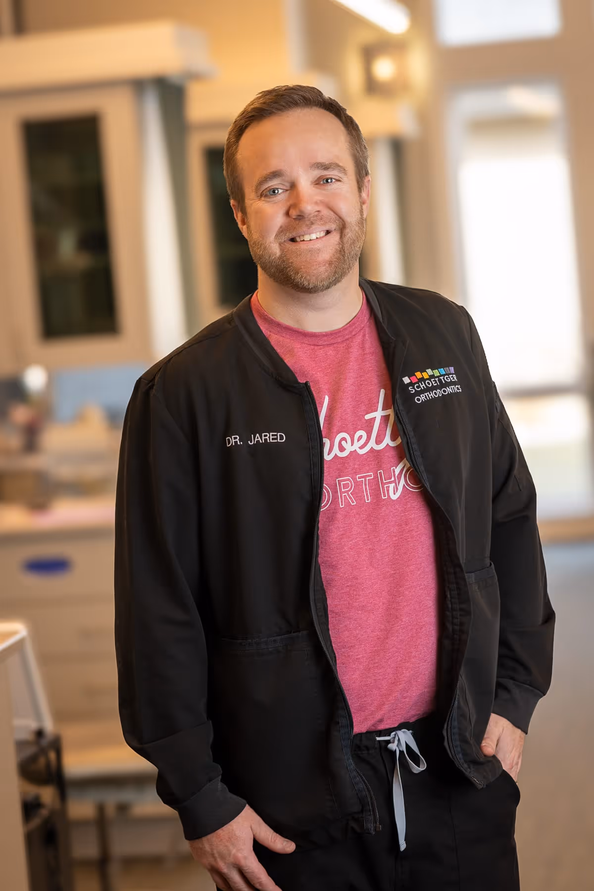 Smiling man wearing a black jacket with 'Dr. Jared' and 'Schoettger Orthodontics' logos, standing in a bright room.