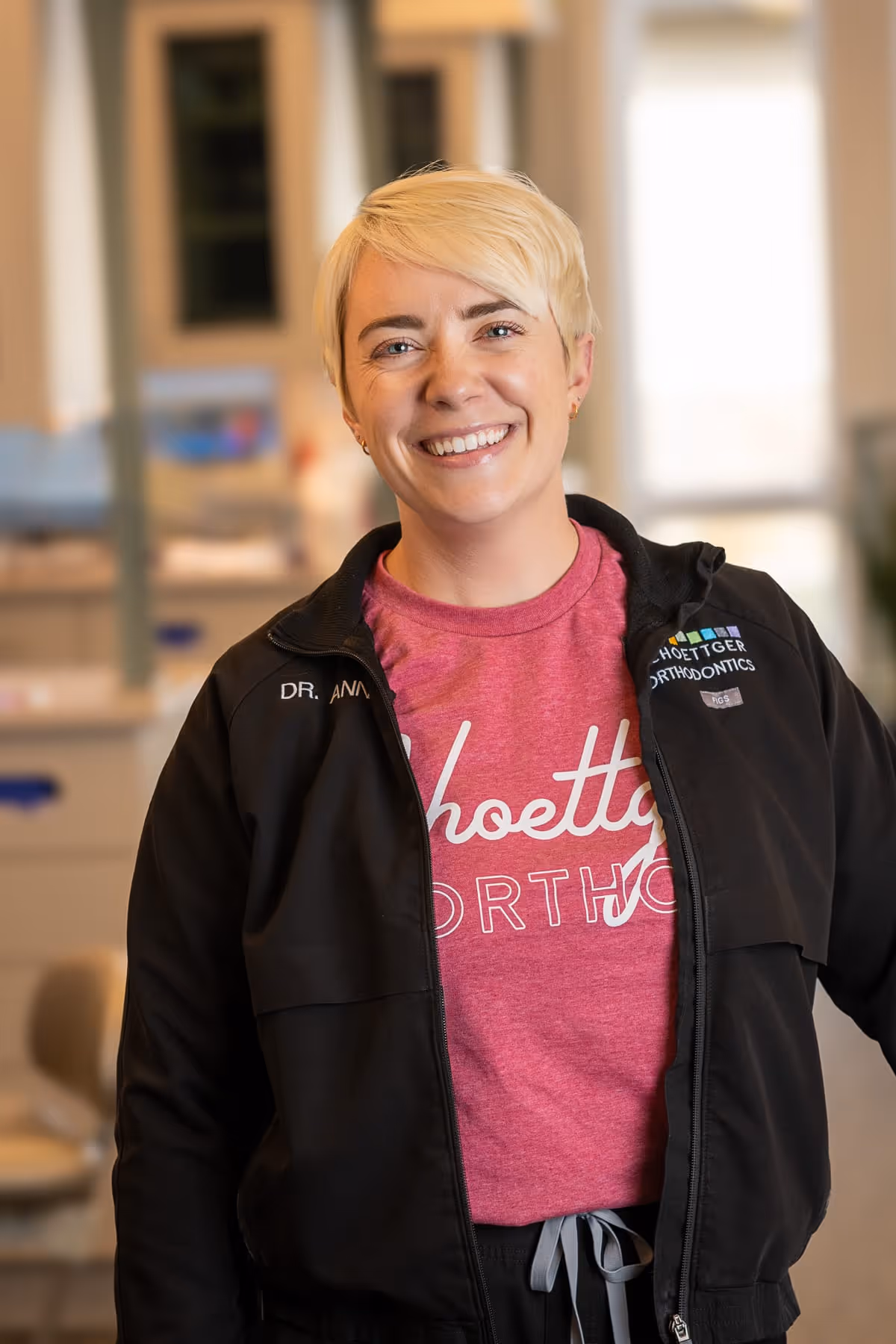 Smiling woman with short blonde hair wearing a pink Schoettger Orthodontics shirt and black jacket indoors.