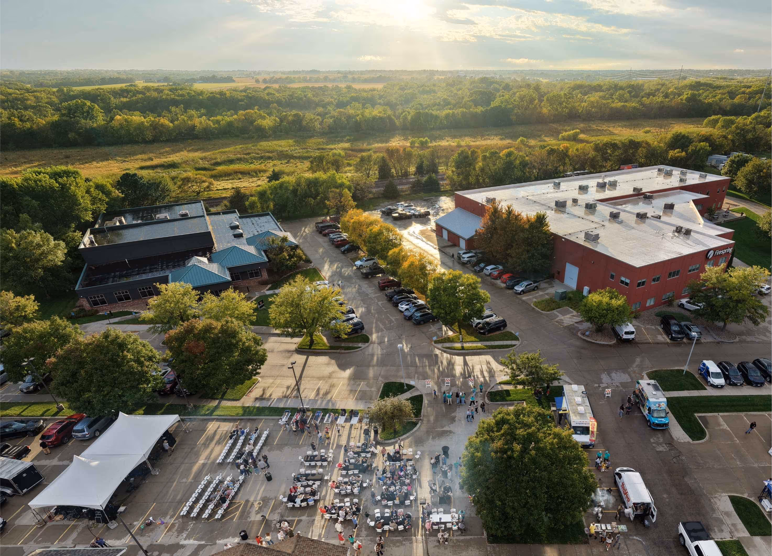 Aerial view of an outdoor event with people seated at tables and rows of chairs in a parking lot, adjacent to two large buildings surrounded by trees and greenery.