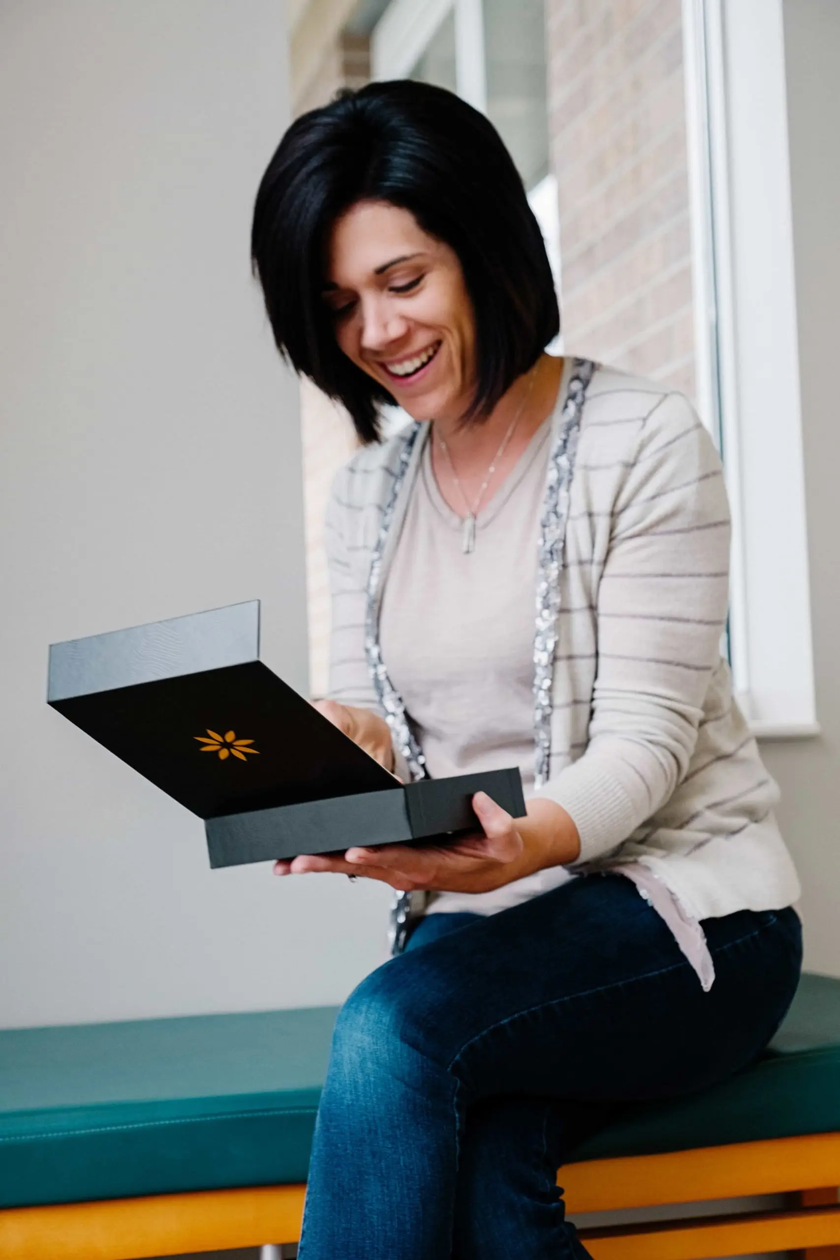 Smiling woman sitting on bench opening a black box with a yellow flower emblem inside.