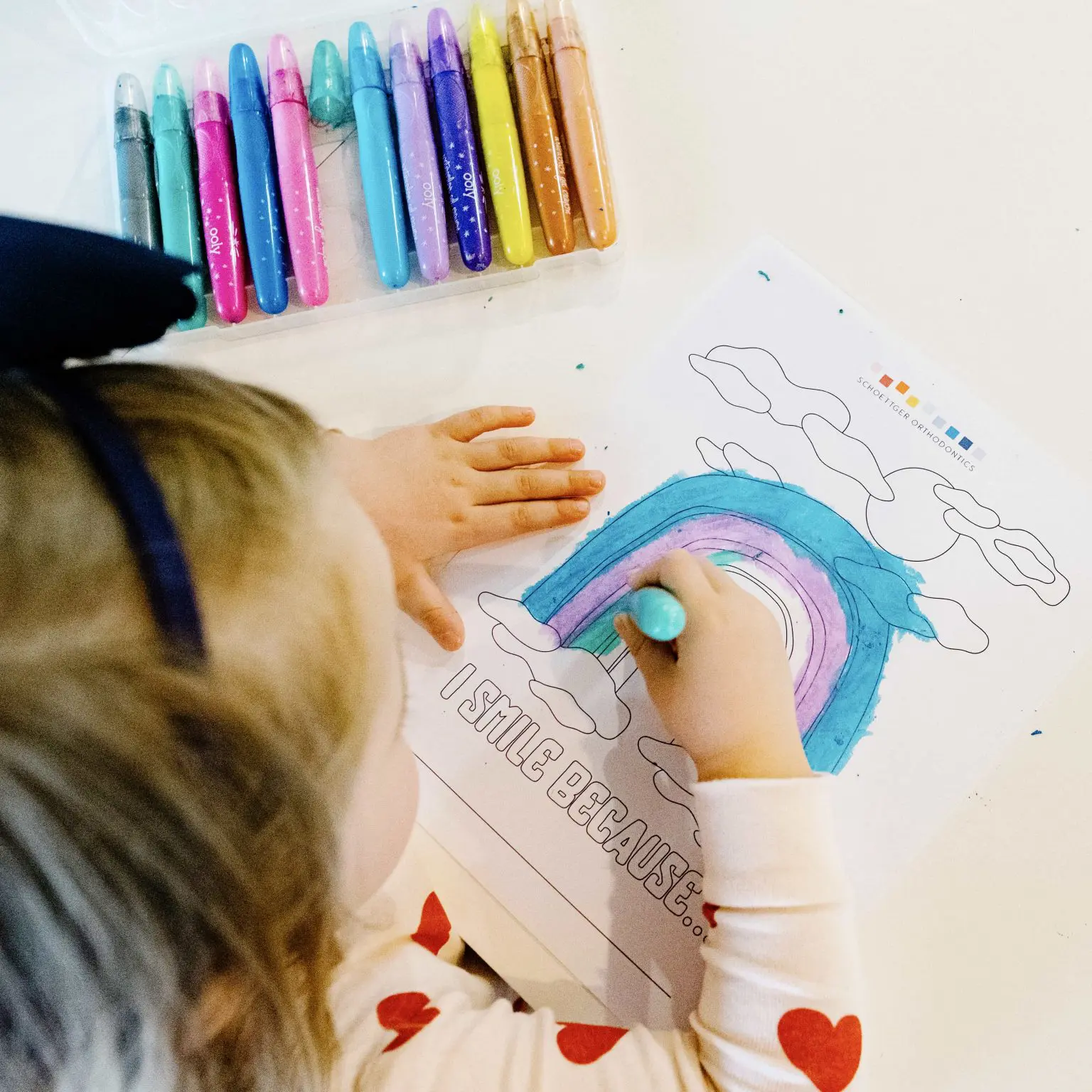 Child coloring a rainbow on a worksheet titled 'I smile because...' with colorful markers nearby.
