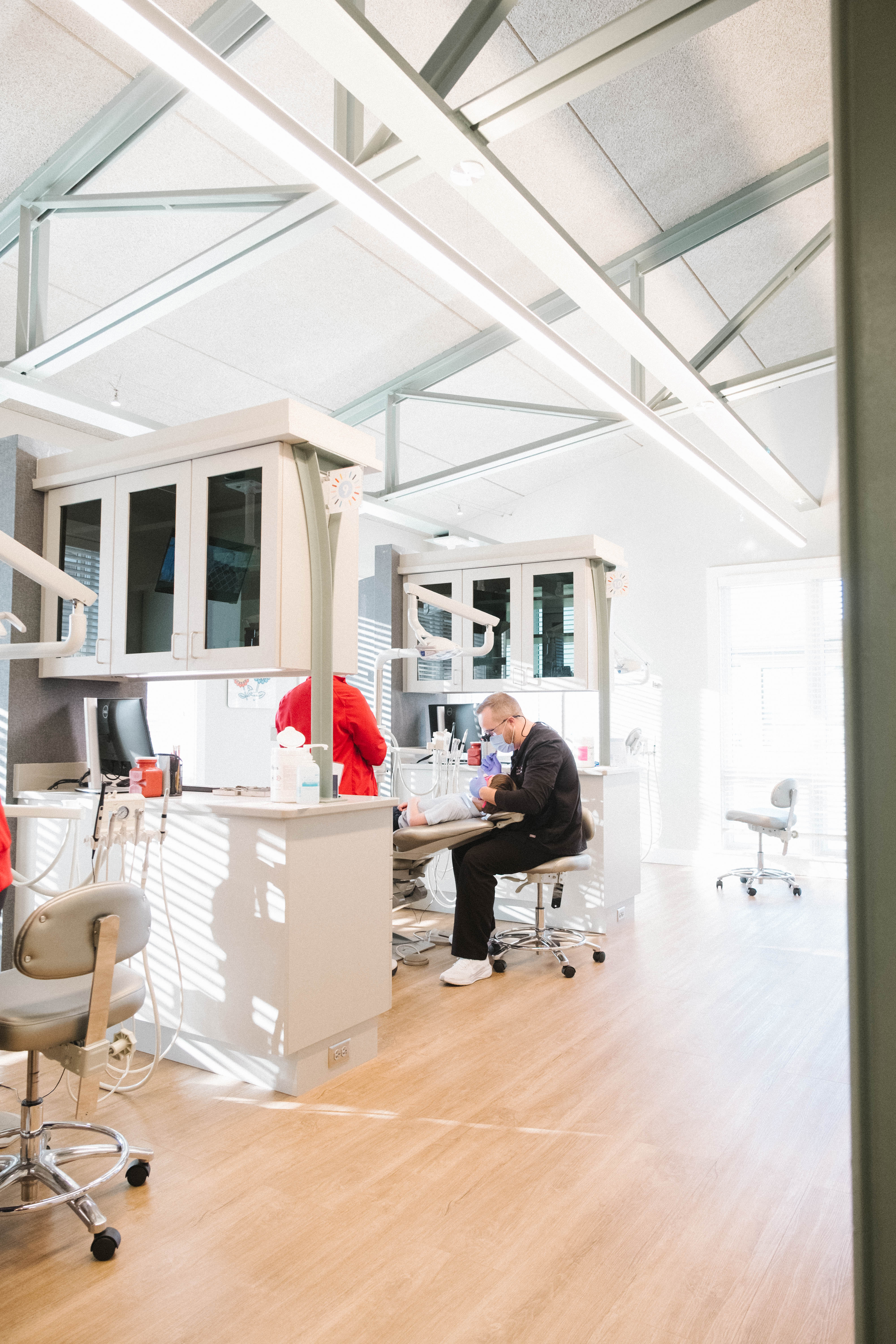Dentist wearing mask treating young patient in modern, bright dental clinic with light wood floors.
