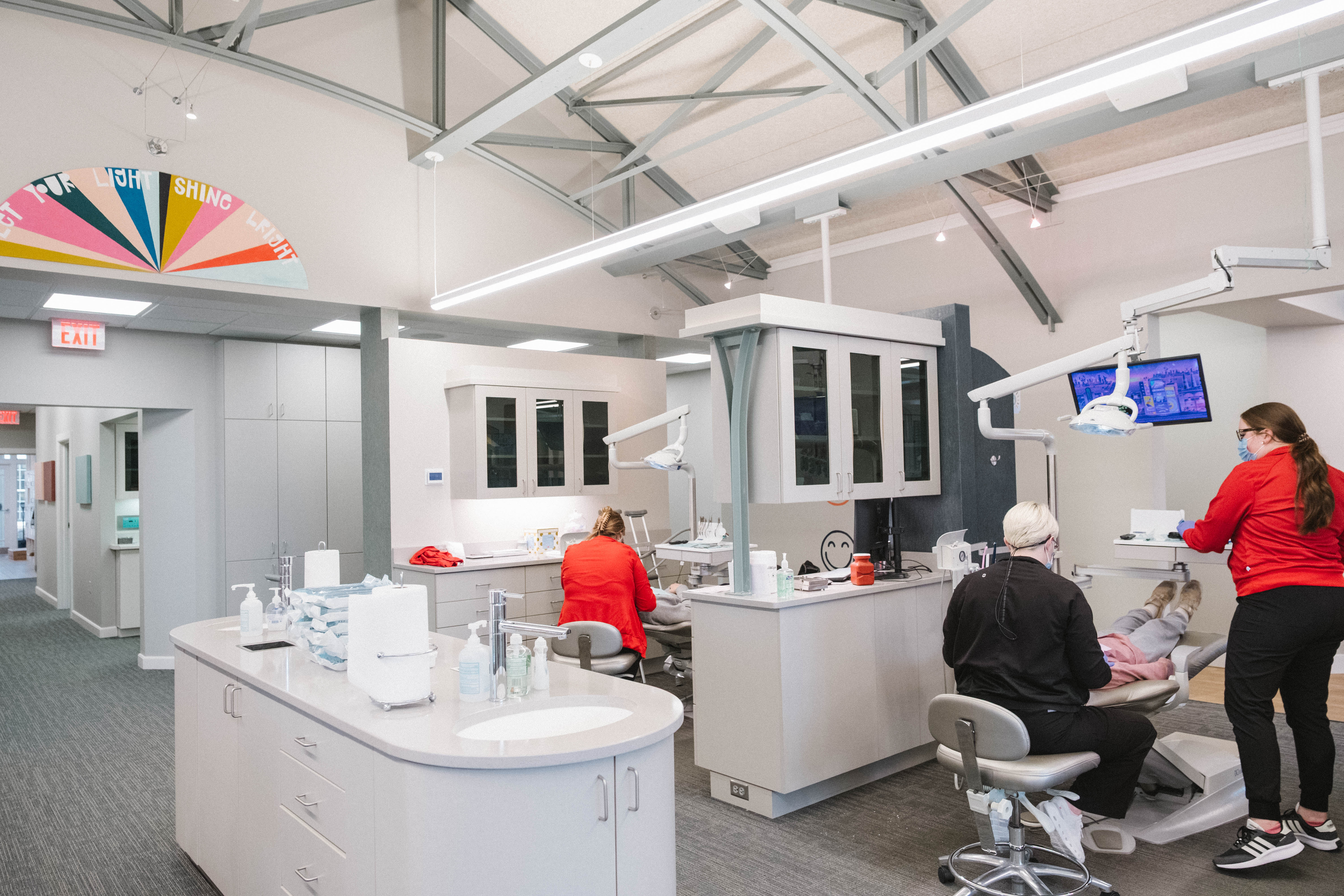 Dental clinic with two patients receiving care from staff in red uniforms wearing masks.