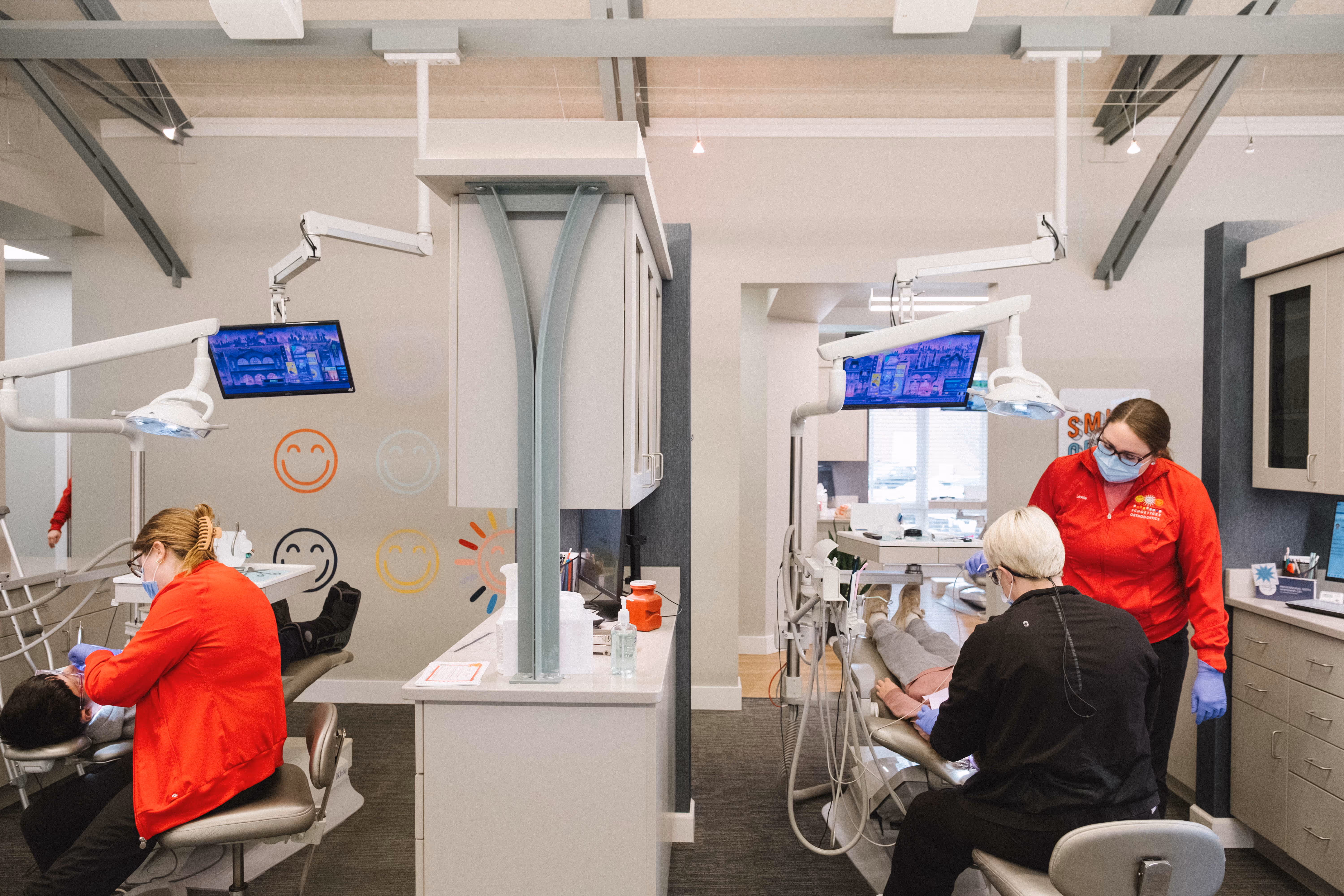 Two dental hygienists in red jackets attending to patients in a bright modern dental clinic.