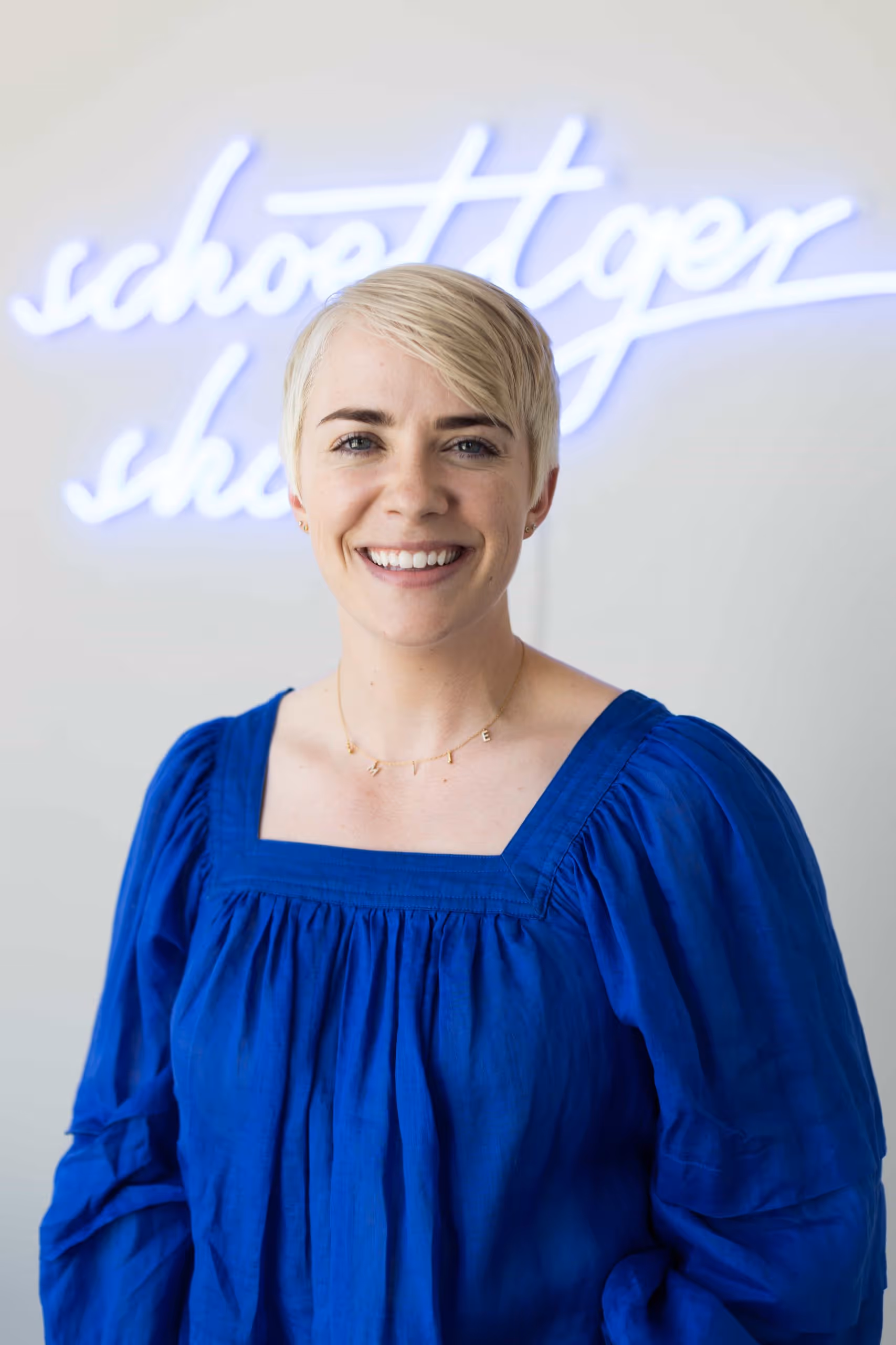 Smiling woman with short blonde hair wearing a blue blouse and gold necklace with letters.
