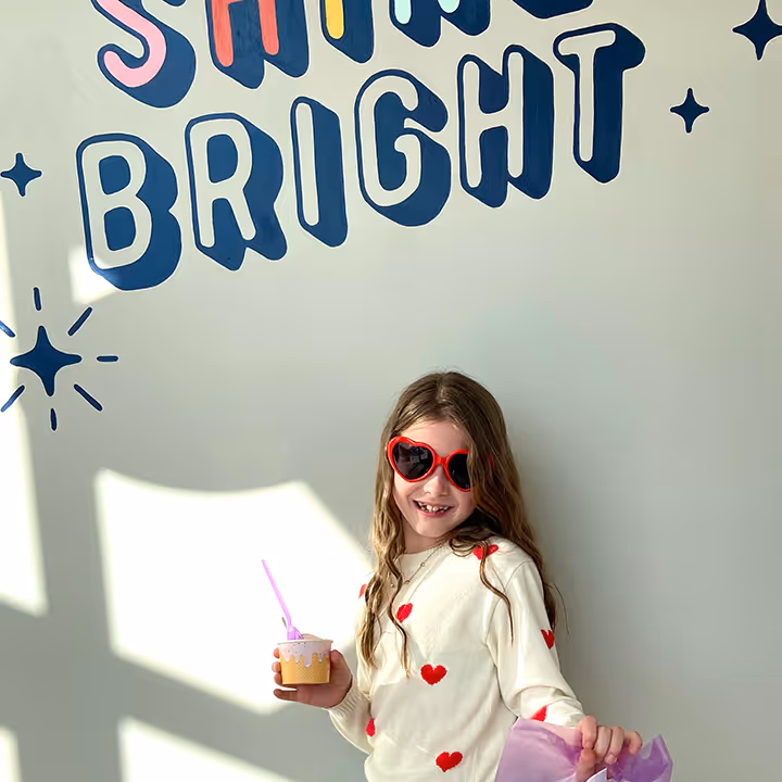 Smiling girl wearing red heart sunglasses and sweater holds a cup with a straw and a purple gift bag.
