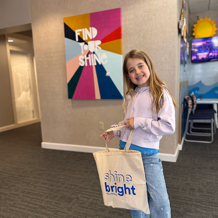 Smiling girl holding a tote bag with 'shine bright' text, colorful 'Find Your Shine' sign behind her.