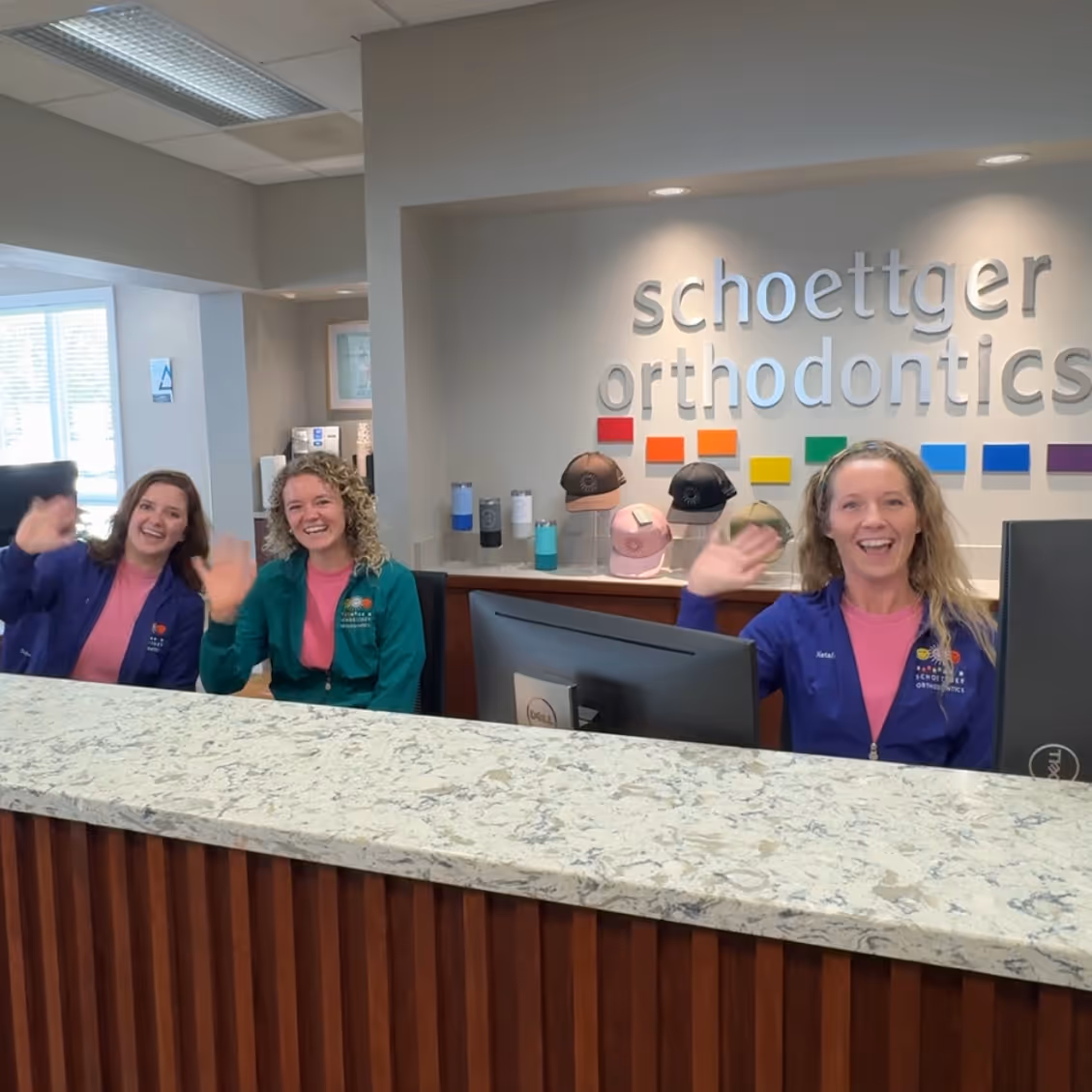 Three smiling women waving behind a reception desk at Schoettger Orthodontics office.