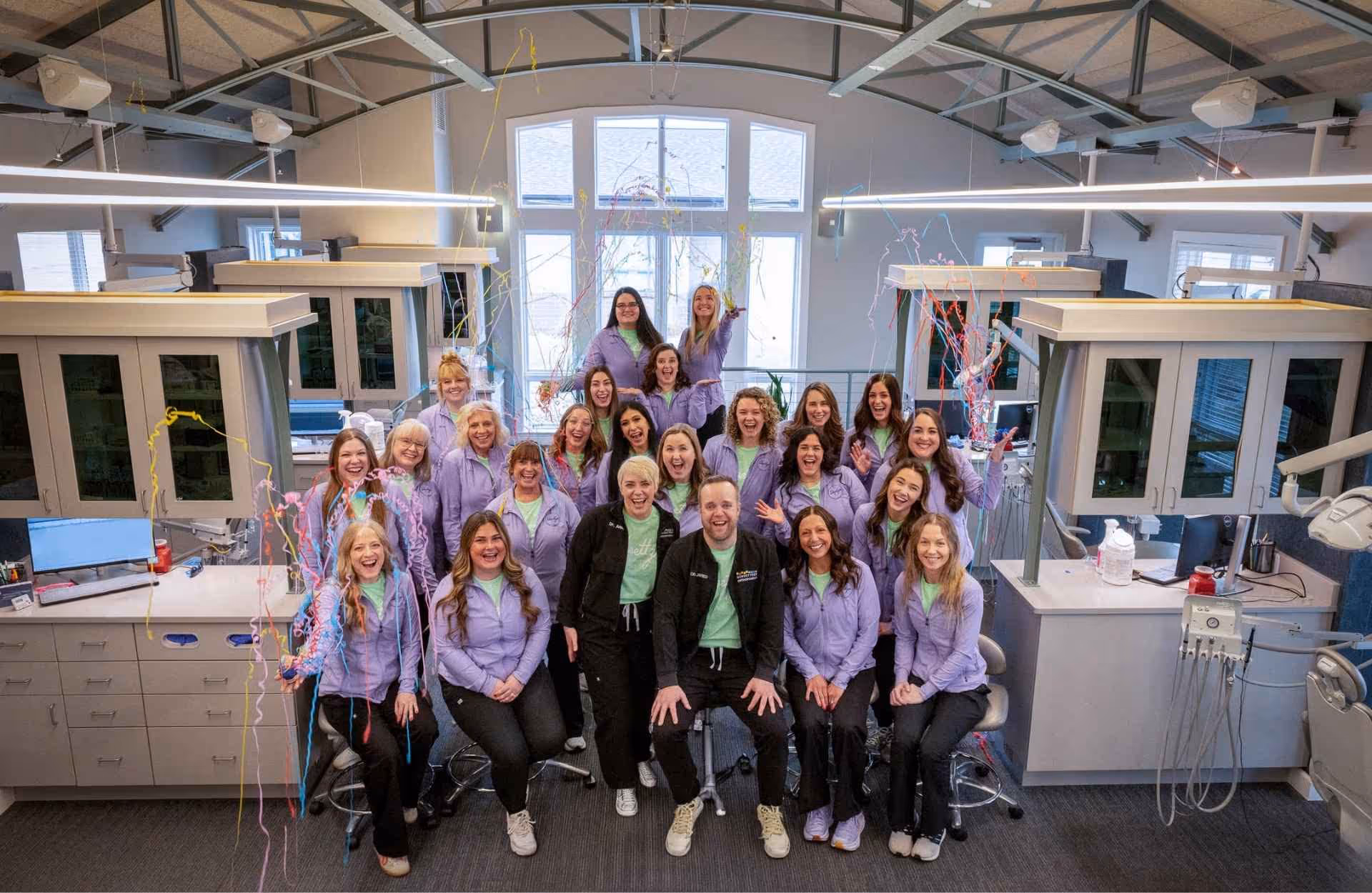 Group of smiling dental office staff in matching purple jackets posing inside a clinic room.