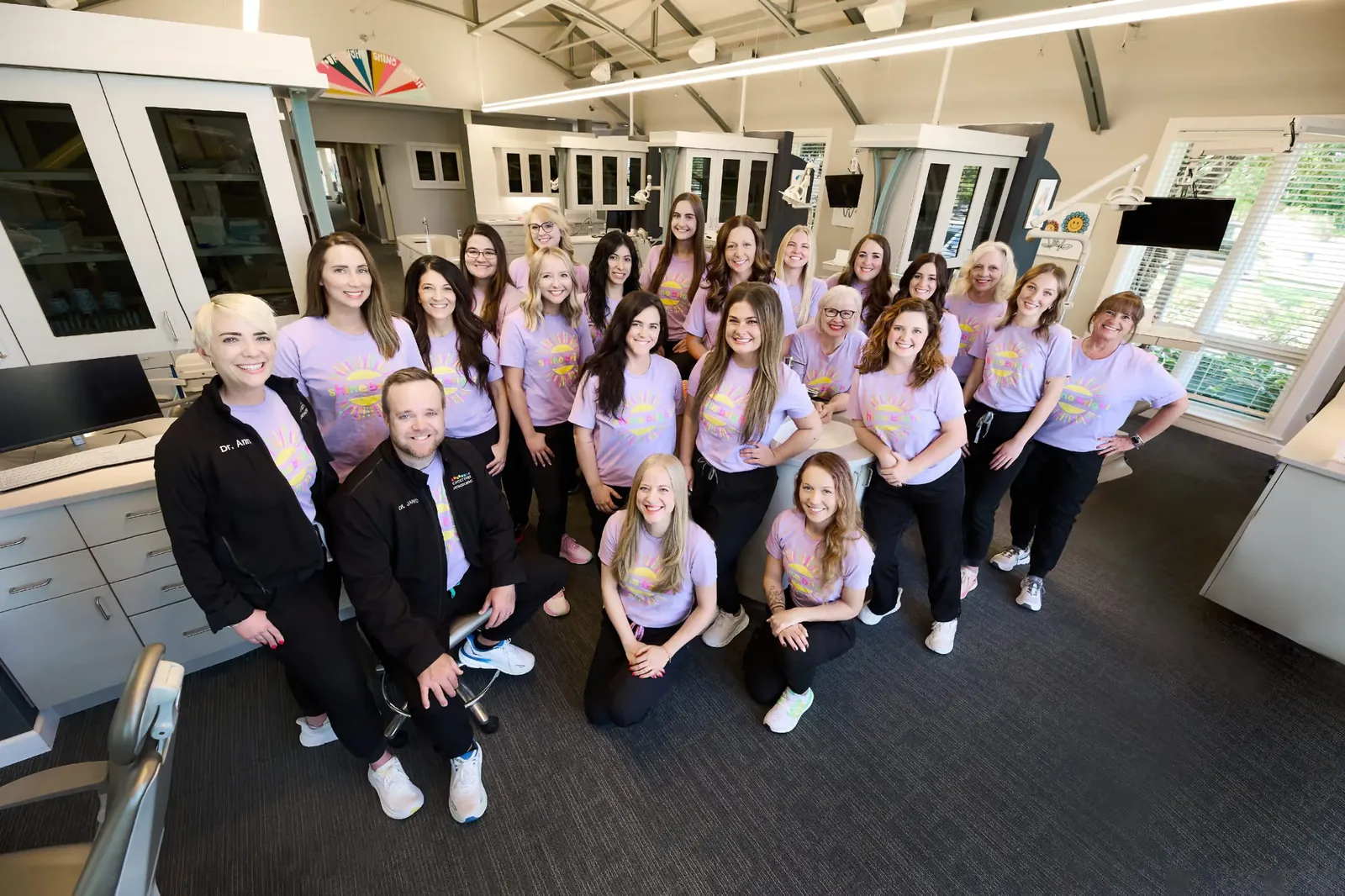 Group of smiling dental office staff wearing matching purple t-shirts posing inside a modern orthodontic clinic.