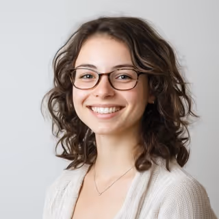 Smiling young woman with curly brown hair and glasses wearing a light sweater and necklace against a plain background.