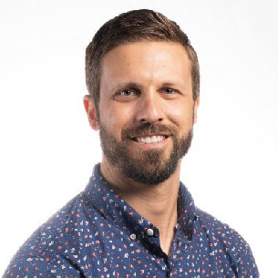 Smiling man with a beard and short hair wearing a dark blue patterned button-up shirt against a white background.