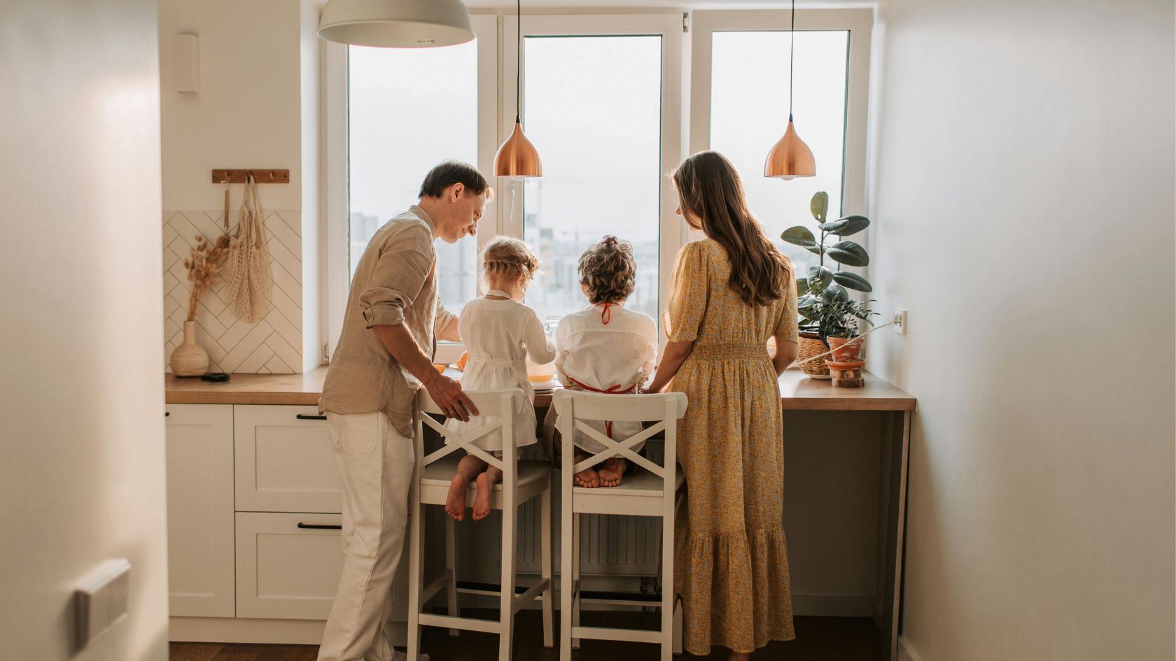 Family of four in a kitchen, two children sitting on stools at a counter with adults standing nearby, preparing food.