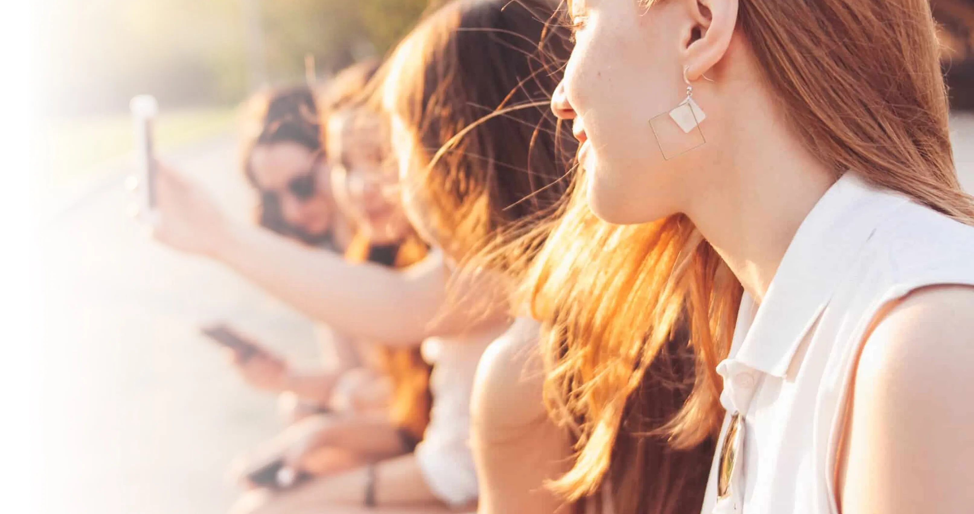 Close-up of a smiling woman with red hair and geometric earrings, with three blurred women in the background holding smartphones.