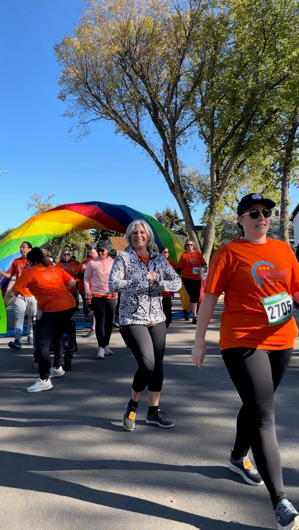 Participants wearing orange shirts walking under a rainbow arch during an outdoor 5K fundraiser event on a sunny day.