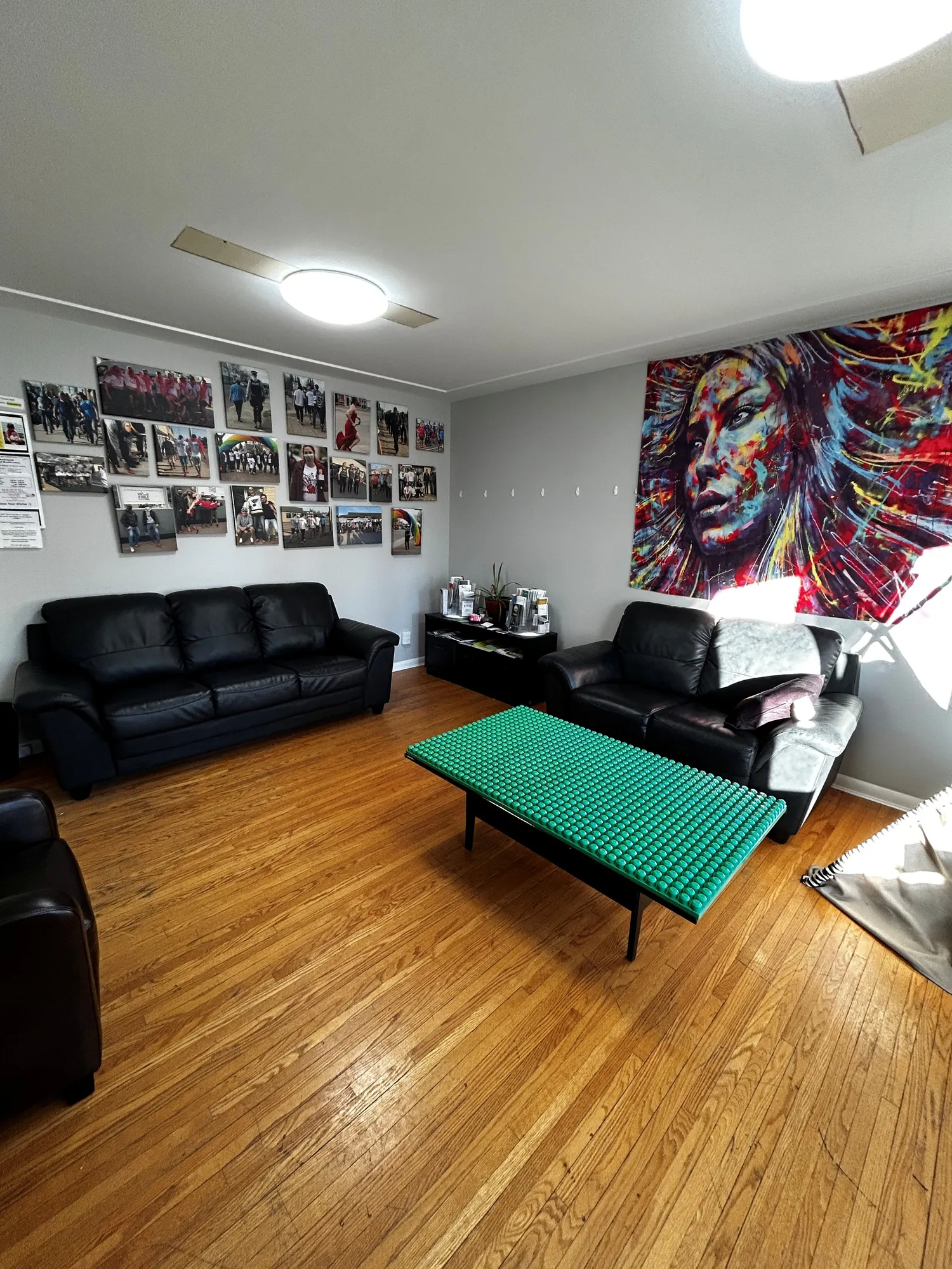 Bright living room with two black leather sofas, a green textured coffee table, photo collage on one wall, and colorful abstract portrait on another.