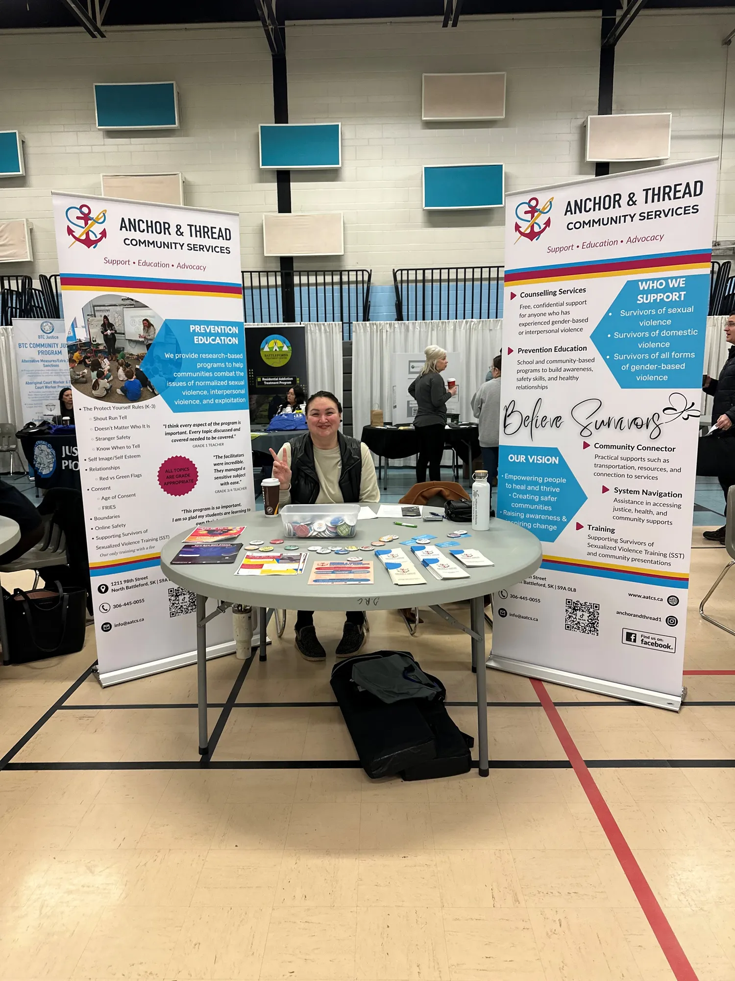 Woman sitting behind a table with informational pamphlets at an Anchor & Thread Community Services booth in a gymnasium.