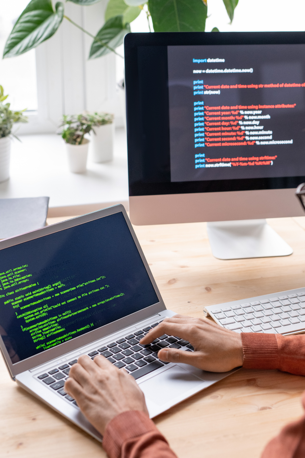 Person tippt auf Laptop-Tastatur mit grünem Java-Code auf dem Bildschirm, im Hintergrund ein Monitor mit Python-Datetime-Code.