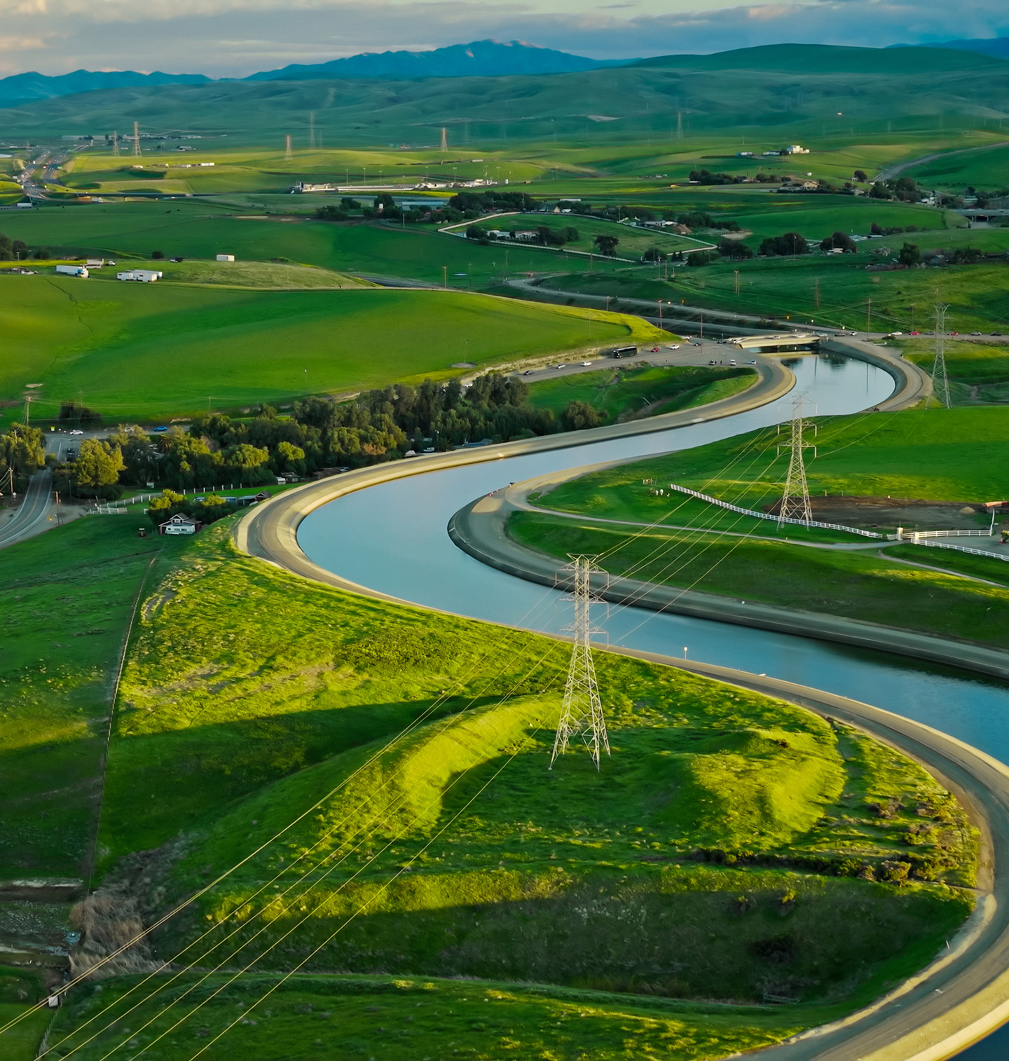 A winding irrigation canal flowing through green agricultural fields with power lines and hills in the background.