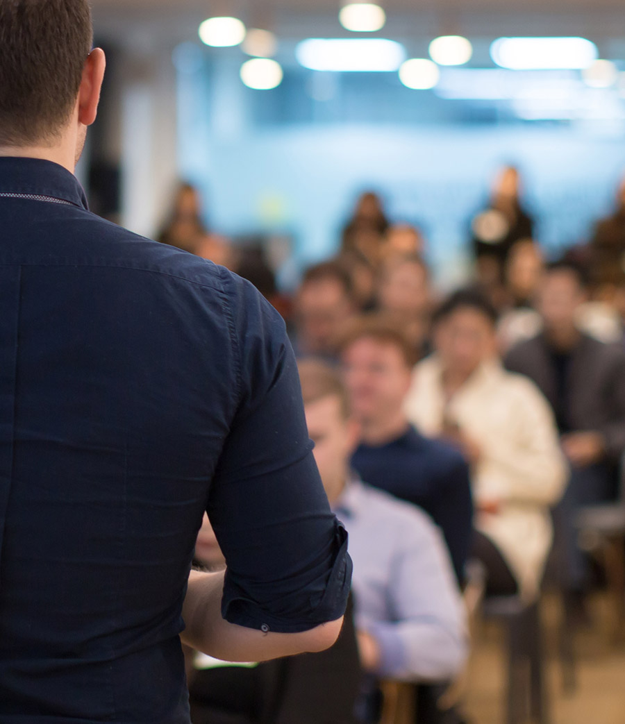Rear view of a man in a dark shirt speaking to a blurred audience seated in a conference room.