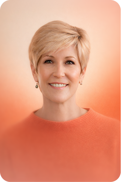 Smiling middle-aged woman with short blonde hair wearing an orange sweater against a soft orange background.