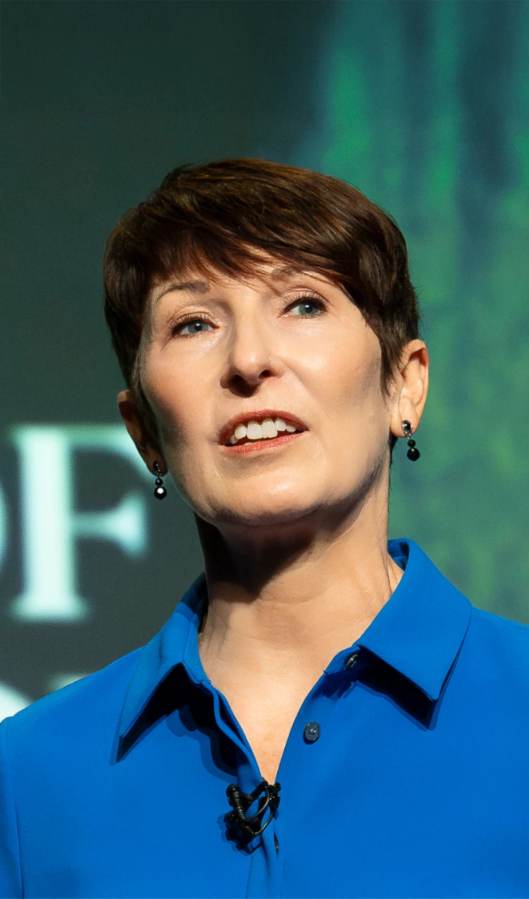 Woman with short brown hair wearing blue shirt and black earrings, speaking with microphone on collar.