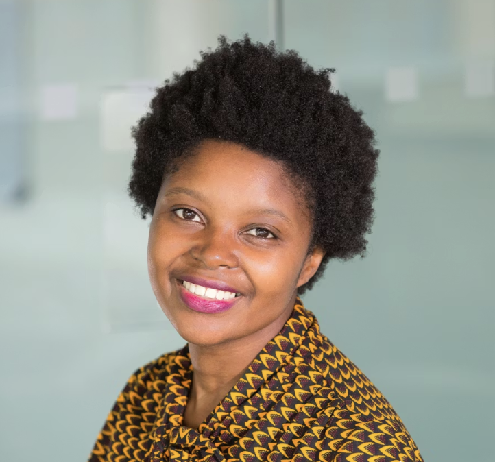 Smiling woman with short curly hair wearing a patterned yellow and black top against a blurred light background.